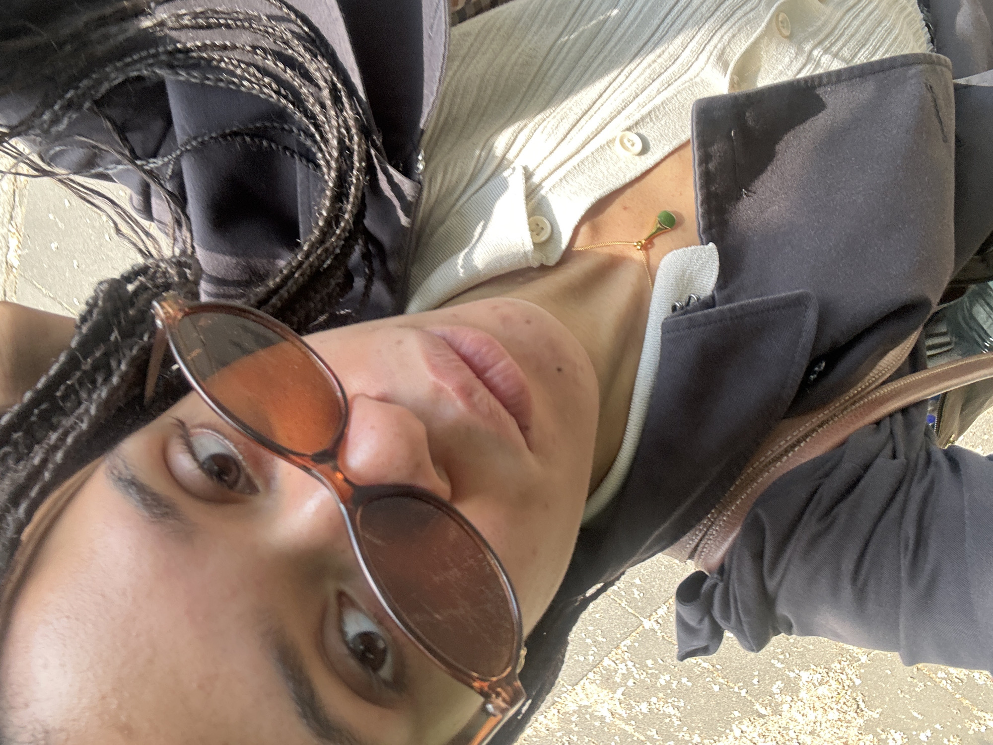 A young woman is pictured in a low-angle shot, seemingly from the ground looking up. She wears sunglasses with amber-tinted lenses and a brown frame. Her hair is styled in braids. She has on a light-colored, button-down cardigan and a dark jacket. A delicate gold necklace with a small, round green pendant is visible at her neck. A brown leather bag is slung across her shoulder. The setting appears to be outdoors, possibly in a city environment, given the textured surface beneath her, which could be pavement.