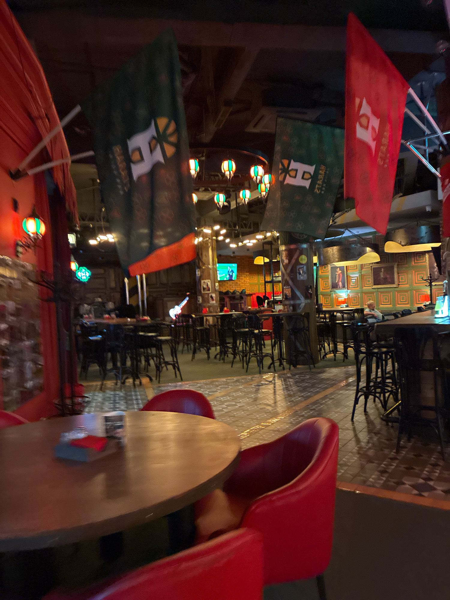 The image shows the interior of a bar or restaurant, with red chairs and tables, and several flags hanging from the ceiling. In the background, there are more tables and chairs, and a stage with a guitar. A person is seated at one of the tables in the background. The overall atmosphere is dimly lit, with decorative lighting adding to the ambiance. The location is identified as a bar in Sochi, Russia.