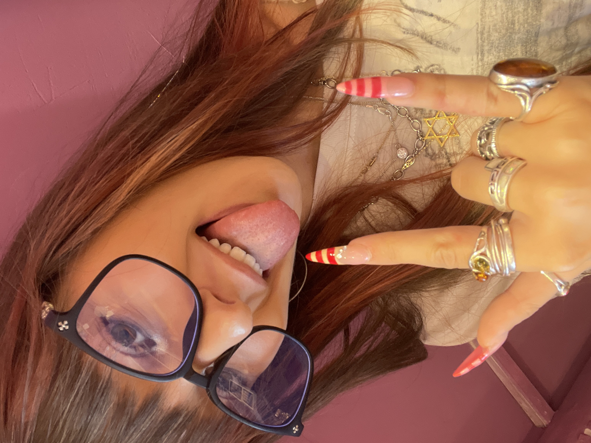 A close-up, high-angle selfie captures a young woman with reddish-brown hair, wearing oversized black glasses with a small metallic embellishment on the left temple. She has her tongue sticking out, and her mouth is open, revealing her teeth. Her right index finger is raised in a "rock on" gesture, and her left hand is making a "shaka" sign. Her nails are long and elaborately decorated; the index finger has red and white stripes, and the thumb nail is a solid bright pink. She is wearing multiple silver rings on her fingers, including one with a large oval amber stone and another with a Star of David pendant necklace. The background is a solid pink wall. The image is taken indoors, with no discernible urban elements or landmarks of Raanana, Israel.