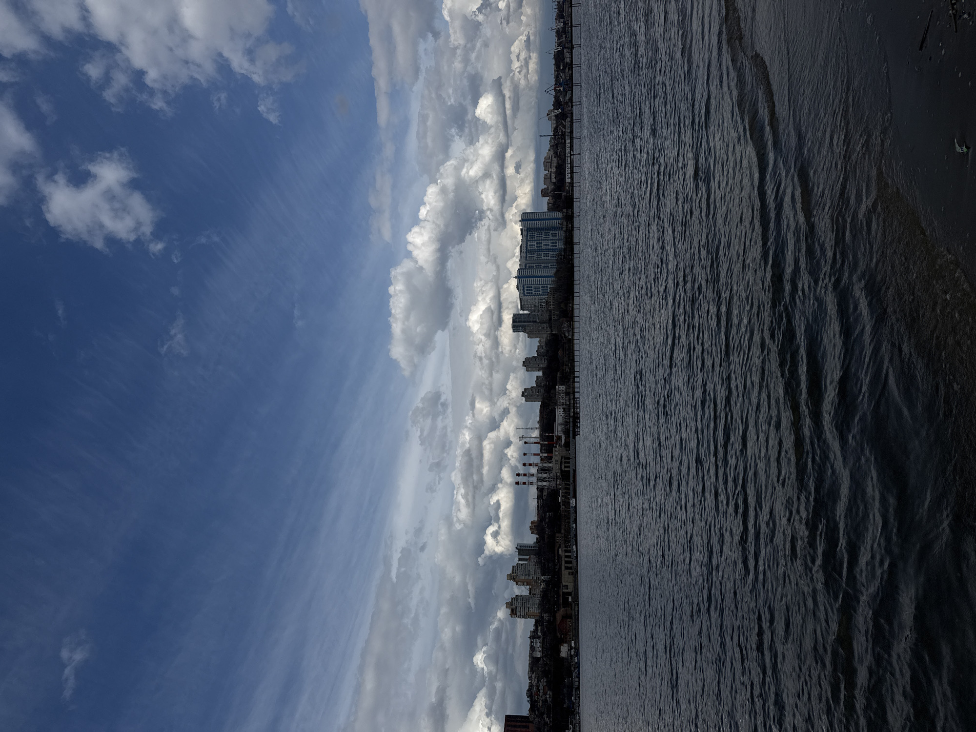 The image depicts a waterfront view in what is likely Rostov-on-Don, Russia. The sky dominates the scene, featuring a mix of blue and striated clouds. Below the horizon, a dark body of water extends toward the viewer, its surface textured by subtle waves. The shoreline is visible on the right. Adjacent to the water, a cityscape rises, including a mix of building styles and sizes. Some buildings appear to be residential high-rises, while others resemble older structures. The perspective is slightly angled, as if taken from a raised position.