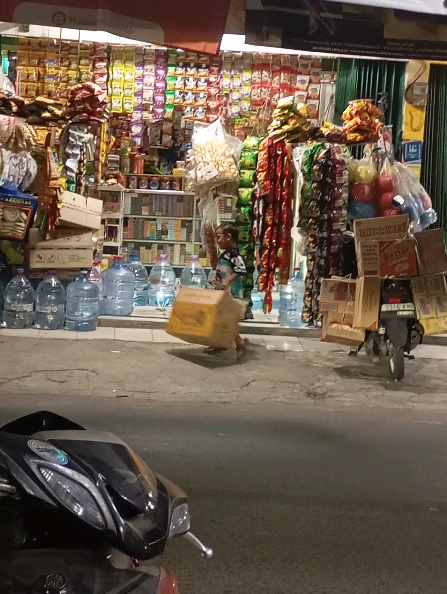 The image depicts a street scene at dusk in Babelan, Indonesia, with a small roadside store in the background. The store is packed with a variety of goods, including snacks, bottled water, and other packaged items displayed on shelves and hanging displays. In the foreground, a young boy, possibly a child laborer, is walking and carrying a large cardboard box. To the right of the boy, a motorcycle is parked, laden with several more cardboard boxes. The street surface appears to be asphalt with some cracks and patches. The lighting suggests it is either early evening or a dimly lit area.