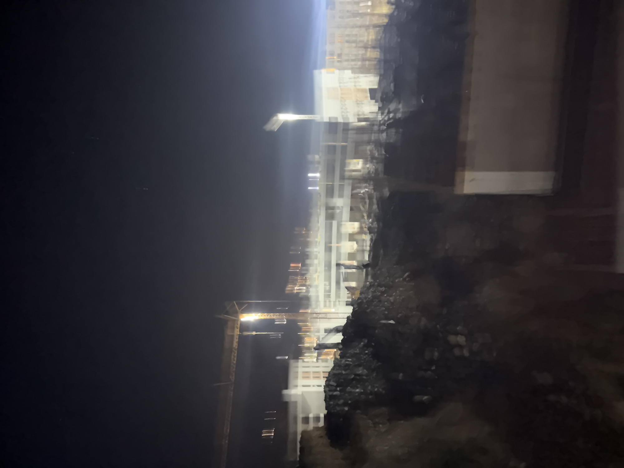 A nighttime aerial view of a construction site in Boreti, Montenegro, shows the blurry outlines of buildings under construction and a large crane. The sky is dark with a faint glow from artificial lights. The foreground is dominated by a pile of dark debris, possibly from demolition or excavation. The blurred nature of the image suggests motion or low light conditions, making it difficult to discern specific details of the construction or any human activity.