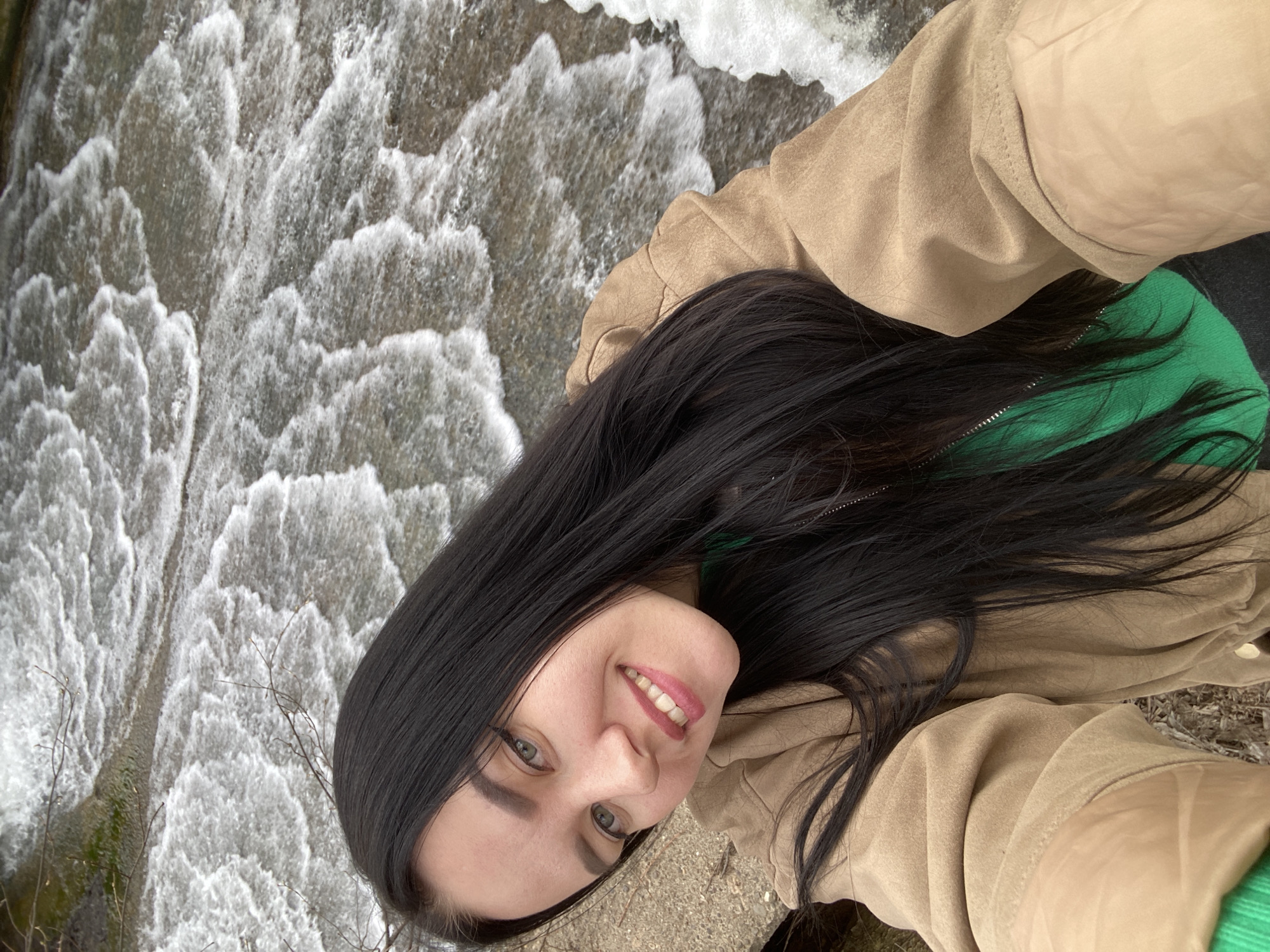A close-up, rotated portrait captures a young woman smiling in front of a cascading body of water. The woman has long, dark, straight hair that falls across her shoulders. She has fair skin, light-colored eyes, and a bright, joyful expression, enhanced by pink lipstick. She is wearing a green top, partially visible, layered under a light brown or tan jacket.
The background is dominated by a large expanse of turbulent, white-foamed water, resembling a waterfall or a dam's spillway, creating a dynamic backdrop. The water appears to be flowing vertically down a textured surface. In the lower left portion of the frame, a few bare tree branches are visible against the moving water. The image conveys a cheerful outdoor moment. No visible text is present. The location context for this image is Фоки, Russia.