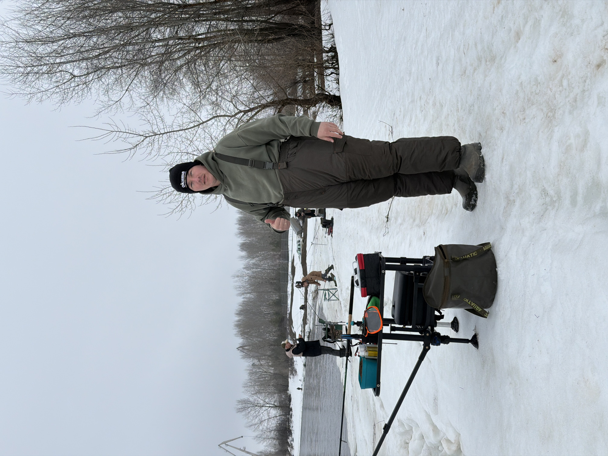 A man wearing a green hooded sweater, dark green overalls, and a black beanie stands in a snowy landscape next to a body of water. He faces the camera with his left arm extended. The beanie has the word "Okuma" printed in white. To the right of the man is a black fishing platform with accessories, including a green bag labeled "AQUATIC." The snowy ground is marked with footprints. In the background, other people are engaged in fishing, along with various fishing equipment. Bare trees are visible along the horizon. Based on the background and other details, the image appears to be taken in Noviy, Russia, where fishing is a common activity.