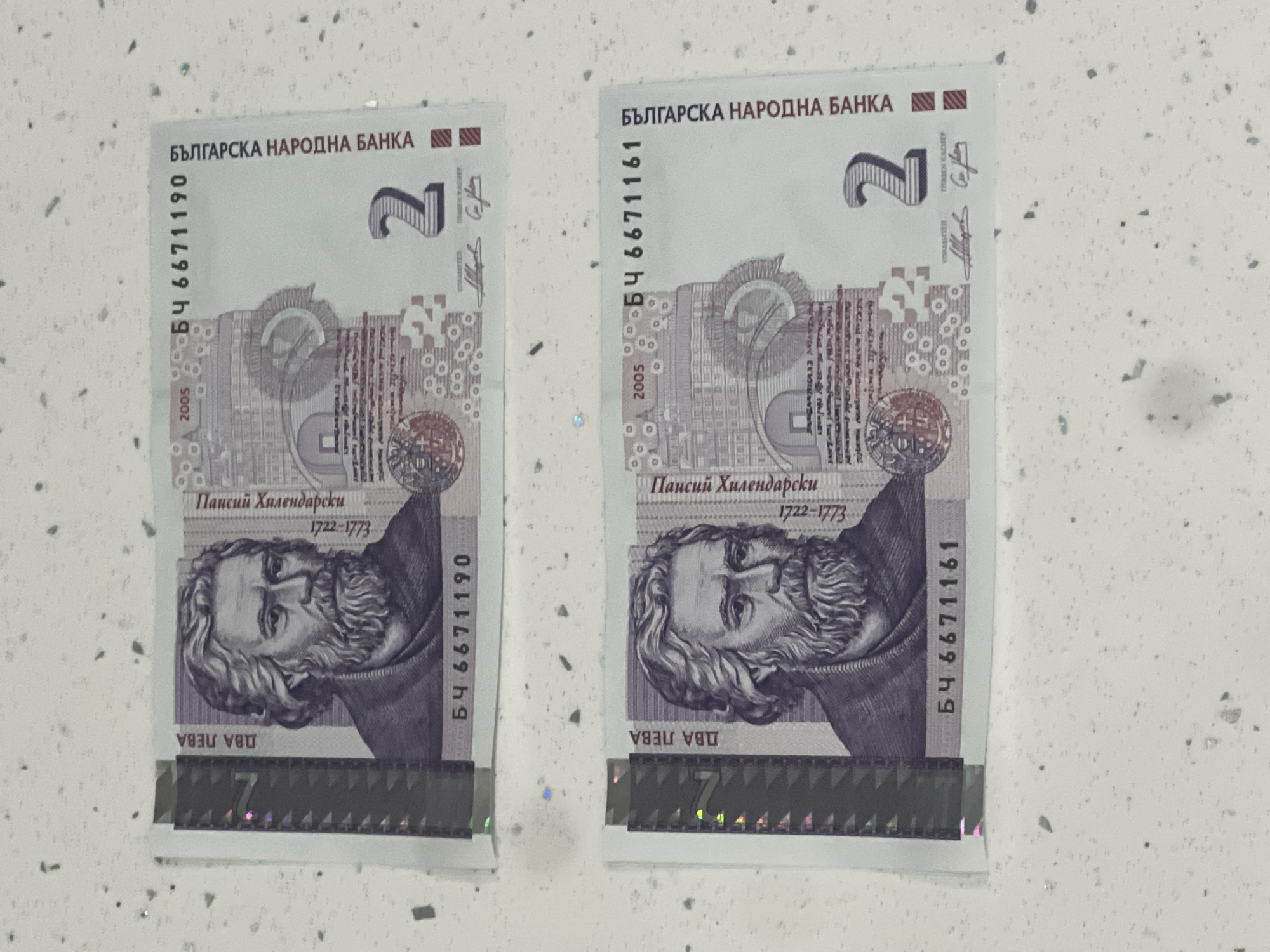 Two Bulgarian 2 lev banknotes are displayed on a speckled white countertop. Each banknote features a portrait of Paisius Hilendarski. The top banknote has the serial number БЧ 6671190 and the year 2005. The bottom banknote has the serial number БЧ 6671161 and the year 2005. Both banknotes are oriented horizontally and are slightly overlapping. The background is a white, speckled surface. There are no people visible in the image. The scene is indoors, likely a domestic setting given the countertop. The lighting appears to be artificial.