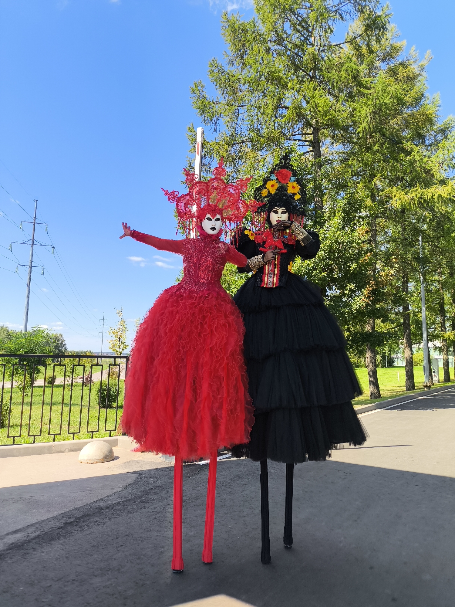Two performers on stilts, dressed in elaborate costumes, pose outdoors under a bright blue sky. The performer on the left wears a vibrant red dress with a full, ruffled skirt and a matching headdress. They have a white mask on their face and their arms are outstretched. The performer on the right is clad in a black dress with a voluminous, tiered skirt and a decorative headpiece adorned with flowers. They are wearing a mask and holding their hands near their face. Behind them, tall green trees form a lush backdrop, and a paved path runs alongside a grassy area enclosed by a fence. Power lines and poles are visible in the distance.