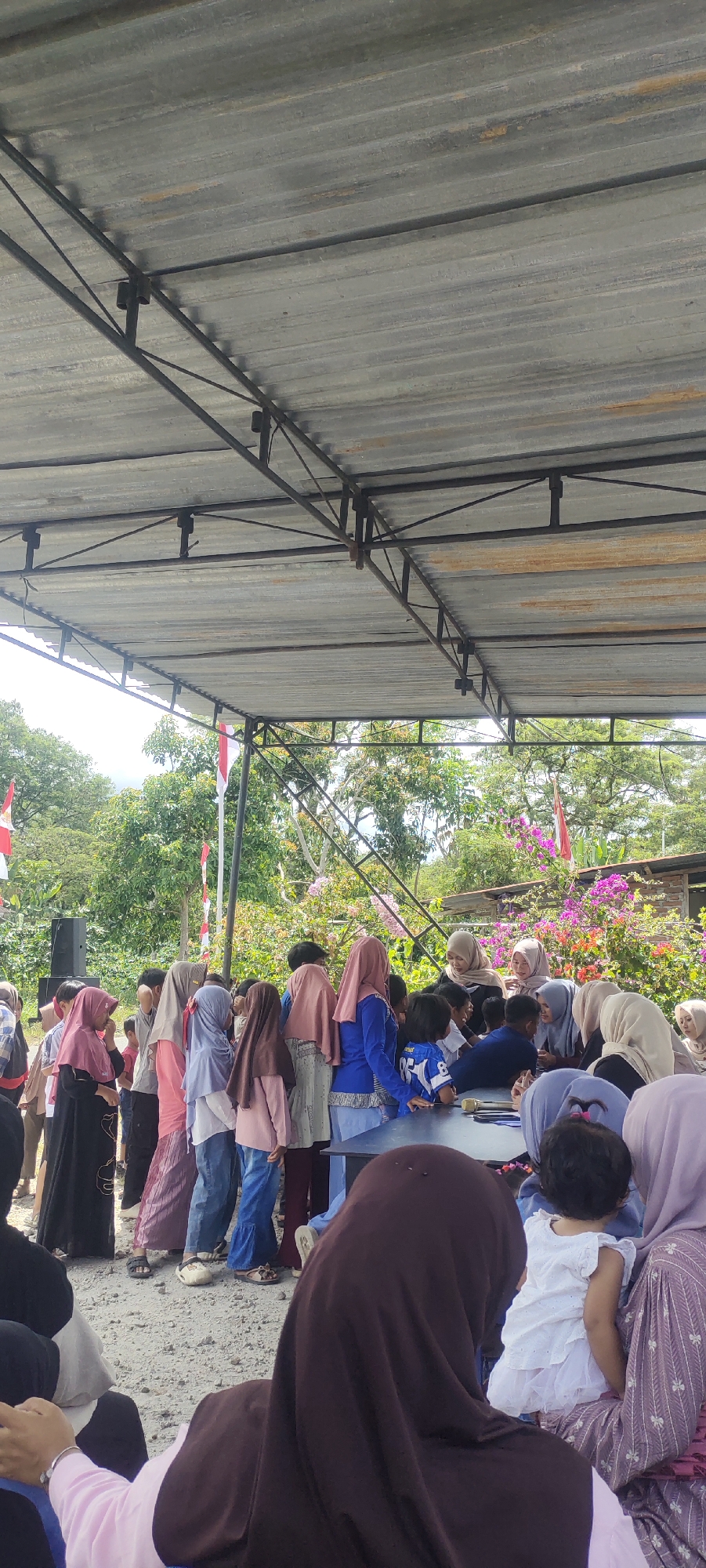 The image depicts an outdoor gathering under a corrugated metal roof supported by a metal frame. In the background, lush green trees and flowering plants are visible, suggesting a natural or semi-rural setting. Indonesian flags, red and white, are mounted on poles and appear to be part of the event's decorations.

The foreground and midground are populated by a group of people, predominantly children and women wearing headscarves and modest clothing. They are gathered around a dark, circular table. Some individuals are looking at the table, while others are interacting with each other, possibly engaging in an activity or a discussion. A black speaker is visible on the left side of the frame. The ground is covered with small stones and gravel. The overall impression is that of a community event or a religious gathering taking place in Simpangantara, Indonesia.