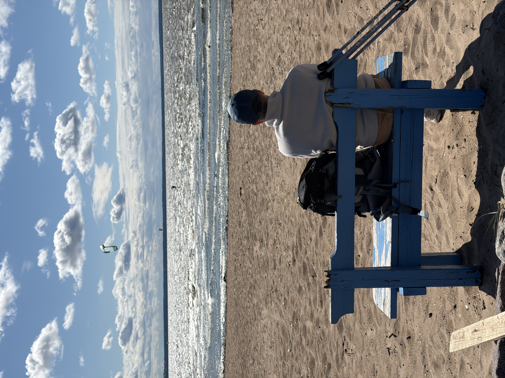 A person sits on a weathered blue wooden bench on a sandy beach, facing the water. The person is wearing a white turtleneck sweater and tan shorts, with a blue baseball cap on their head. A black backpack rests on the bench beside them, and a pair of ski poles are propped up against the back of the bench to their right. The beach is composed of light brown sand with scattered debris. In the distance, the choppy, sun-dazzled water stretches to the horizon, meeting a sky filled with scattered white clouds. Several kitesurfers are visible on the water, and one kite is prominent in the sky. The scene is likely located on one of the beaches of Kronstadt, Russia, known for its maritime history and proximity to the Gulf of Finland.