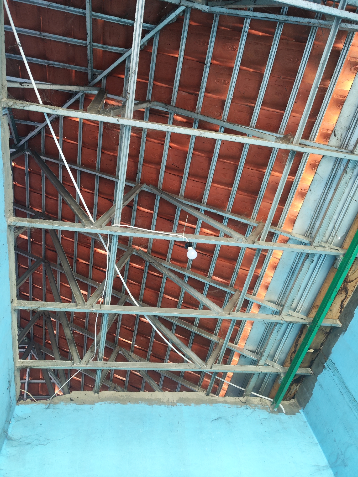 The image depicts an unfinished or under-construction roof structure with visible terracotta tiles and a metal frame. The interior walls are painted light blue. A single light bulb hangs from a white wire. The scene appears to be in a residential area, possibly in Sragen, Indonesia, given the architectural style and materials. There are no people visible in the image.