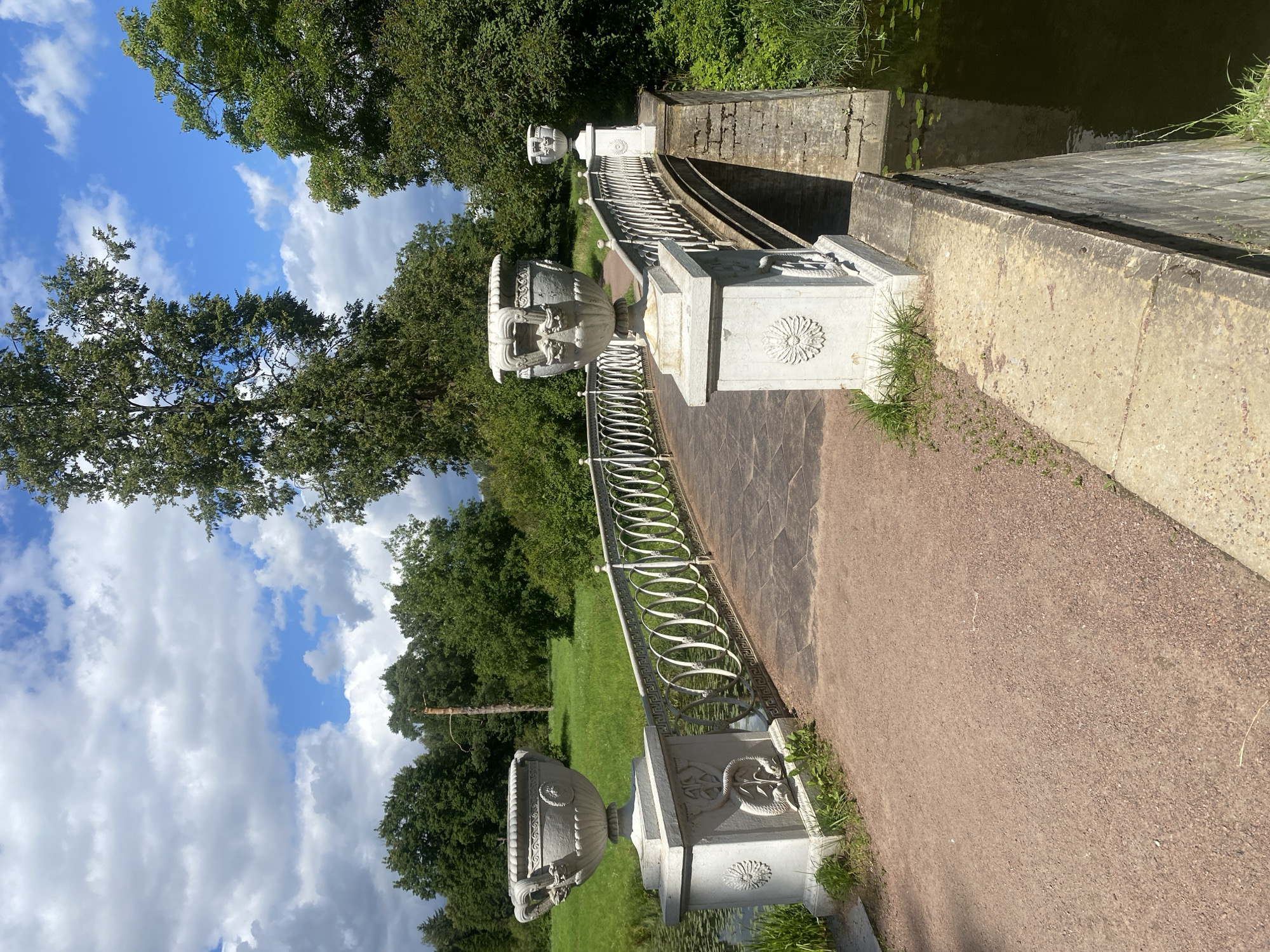 The image shows a stone bridge with an ornate white railing crossing a body of water in Pavlovsk, Russia. Two large decorative urns are placed on pedestals at the entrance of the bridge. The bridge is paved with stone tiles and a sandy path leads up to it. Lush green trees and grass surround the bridge, and the sky is partly cloudy with patches of blue. The scene depicts a picturesque park setting.