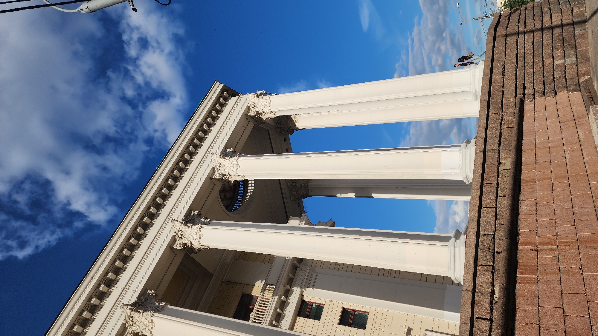 The image shows an upward-angled view of a building with large, white, classical-style columns against a backdrop of a blue sky with scattered clouds. The building facade is light-colored with architectural detailing. To the right of the columns, a set of brick steps is visible. Two individuals are on the stairs.
The location is likely Moscow, Russia.