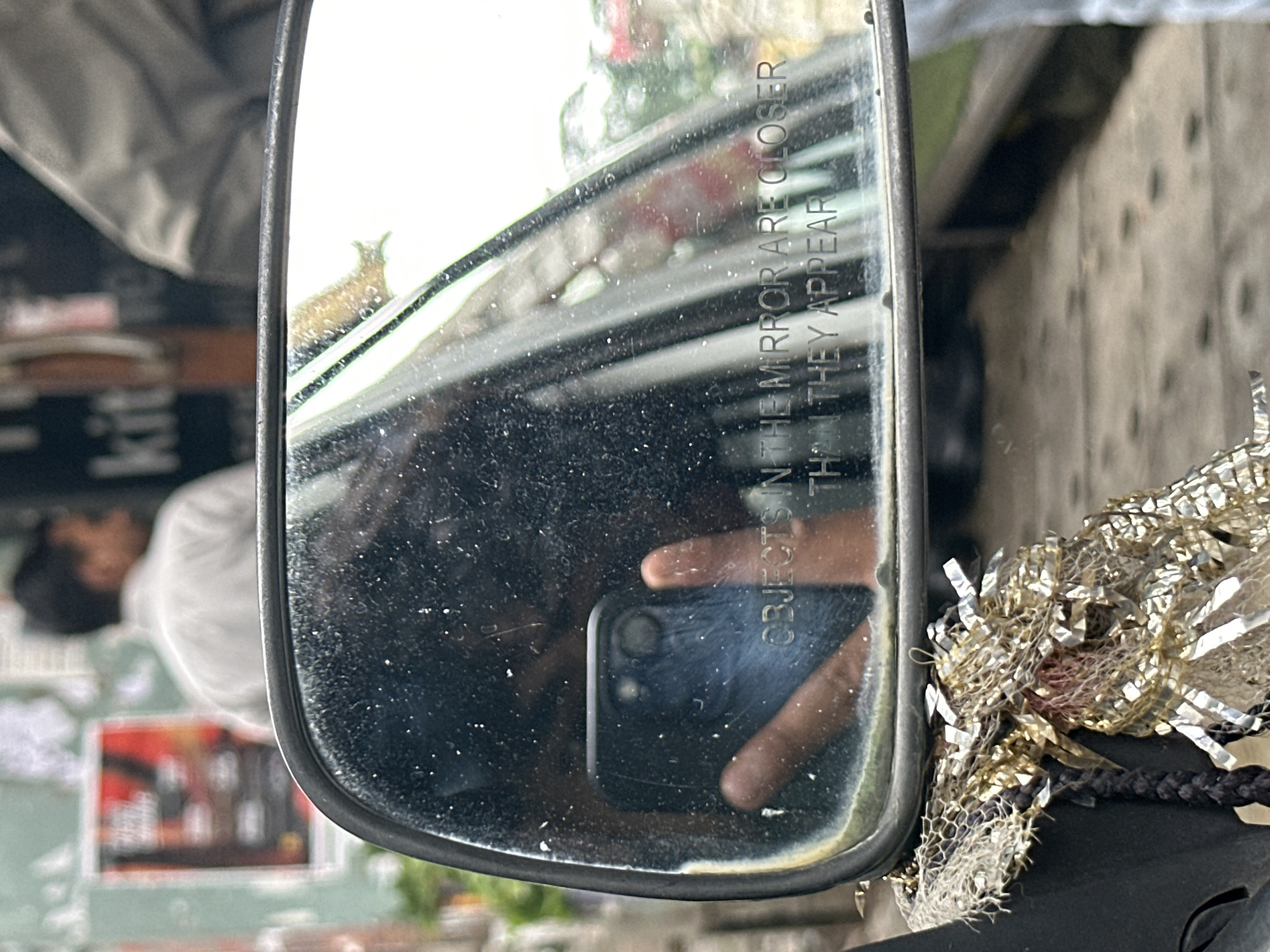 In this close-up shot, a side-view mirror of a vehicle is the primary focus, reflecting a scene in motion. The mirror's surface is speckled with dust and debris, obscuring some details. In the reflection, a hand holds a dark-colored smartphone, positioned to capture the mirror's view. The text "OBJECTS IN THE MIRROR ARE CLOSER THAN THEY APPEAR" is clearly visible on the mirror's surface, a common warning on such mirrors.
The background of the reflection shows a busy street scene. A man in a light-colored shirt is visible in the distance, his back turned to the camera. Behind him, a poster with red and black elements is partially visible on a wall. To the right, another person, seen from the shoulder down, wears a grey jacket. The street itself appears to be paved with cobblestones or similar textured material.
In the foreground, a decorative element, likely attached to the vehicle, dangles in front of the mirror. It is composed of interwoven mesh and shiny, metallic-looking ribbons, giving it a festive or ornamental appearance. The overall lighting suggests an overcast day.