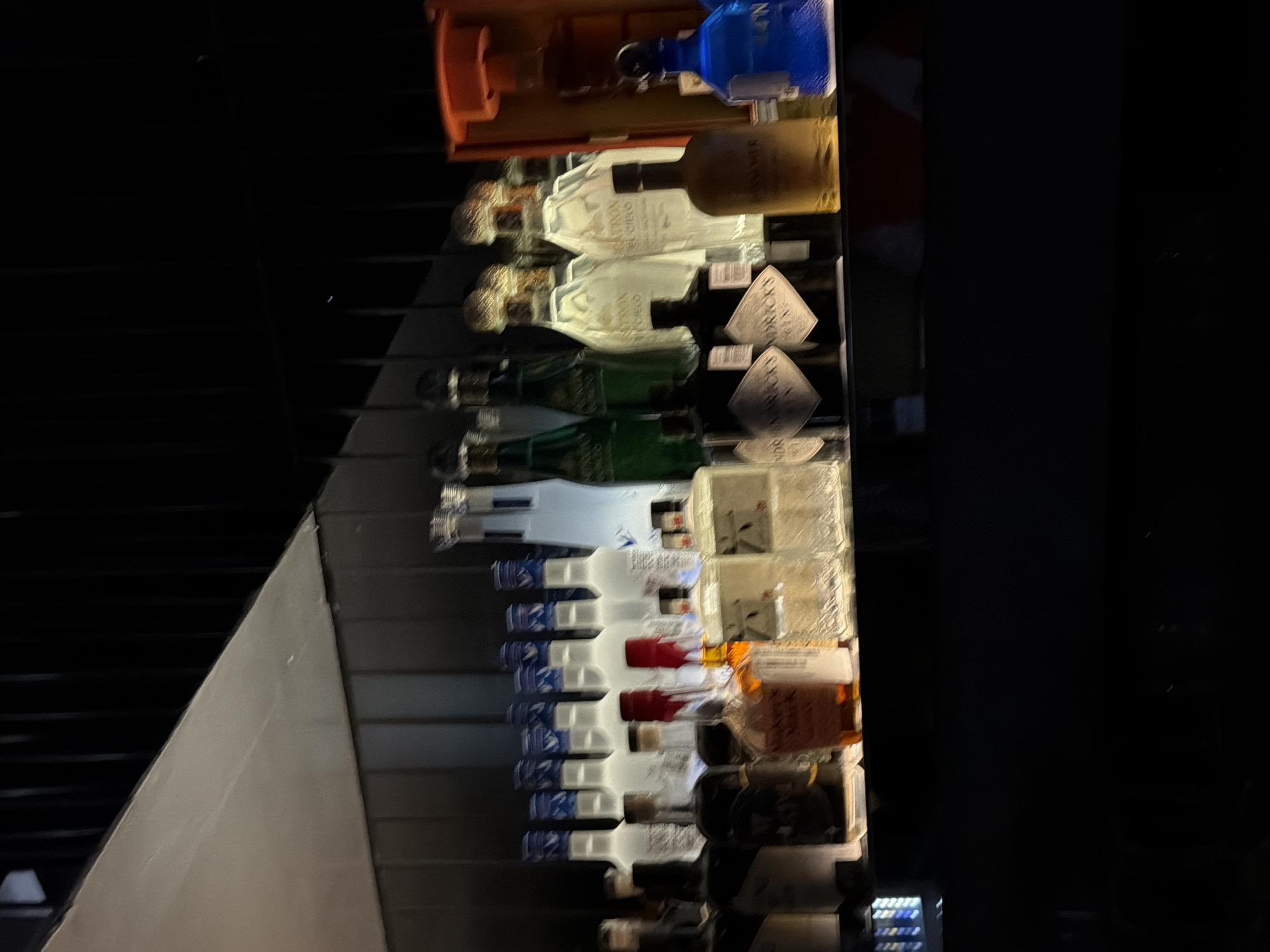 A static, dimly lit indoor scene captures a close-up of premium liquor bottles displayed on an illuminated bar shelf, located within an establishment in New Delhi, India. The bottles are arranged in rows, primarily illuminated from behind or below, creating distinct glows through the glass.
Key identifiable spirits include:
* A row of multiple frosted or clear vodka bottles with blue caps, consistent with Grey Goose, occupying the upper left section.
* A Maker's Mark bourbon bottle, recognizable by its amber liquid and red wax-sealed cap, positioned centrally among other brown spirits.
* Two dark, square-shouldered Hendrick's Gin bottles, featuring their characteristic diamond-shaped labels, situated in the lower-middle right.
* Several tall, clear bottles with intricate stoppers and light-colored liquid, identified as Patrón Tequila, with "PATRON EL CIELO" visible on one label, present in the upper right.
Additional bottles include two green-hued spirits, various clear and amber-colored spirits, and a single bright blue bottle on the far right. A partial view of an orange-brown wooden container is visible on the rightmost edge. The background consists of dark, vertically oriented elements, likely a paneled wall or decorative bars. An angled, light-colored structural surface is visible in the upper left corner. No individuals are present, and no actions or interactions are depicted. The composition focuses solely on the arrangement and illumination of the bottled spirits.