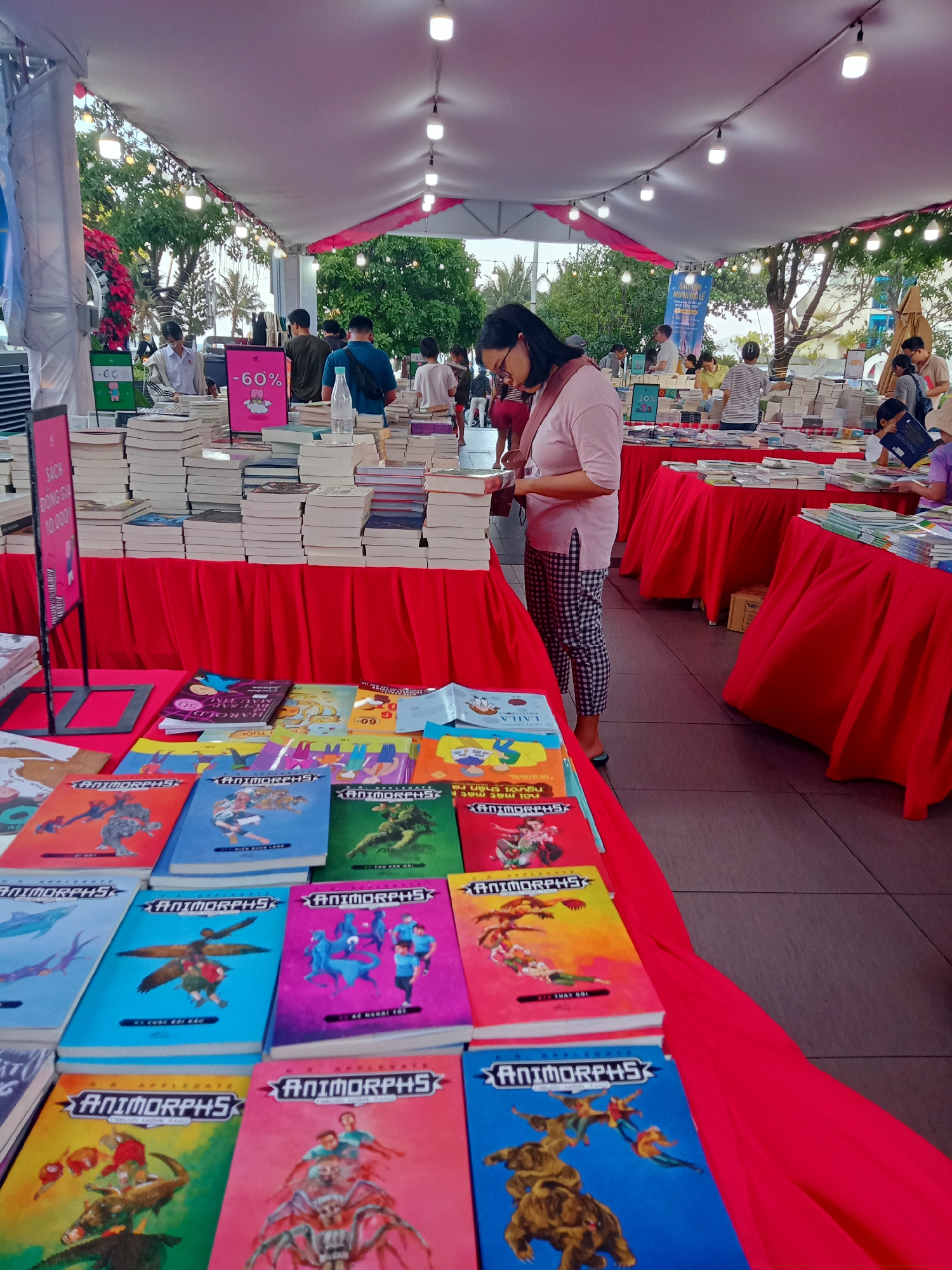 The photograph depicts a bustling book fair scene, seemingly taking place outdoors under a tent or canopy, possibly in the city of Nha Trang Ward, Vietnam. The scene is filled with tables draped in bright red fabric, laden with stacks of books. In the foreground, a table displays numerous copies of the "Animorphs" book series with covers in various bright colors. A woman stands at one of the tables, dressed in a light pink top and checkered pants, engrossed in examining a book. In the background, other individuals are perusing the book selections. Signage indicates discounts such as "60%" and prices such as "10,000", likely referring to the local currency. The environment is bright and airy, suggesting an outdoor setting with trees and other elements of urban greenery visible in the distance. Overhead, strings of lights hang beneath the tent, adding to the fair's festive atmosphere.
