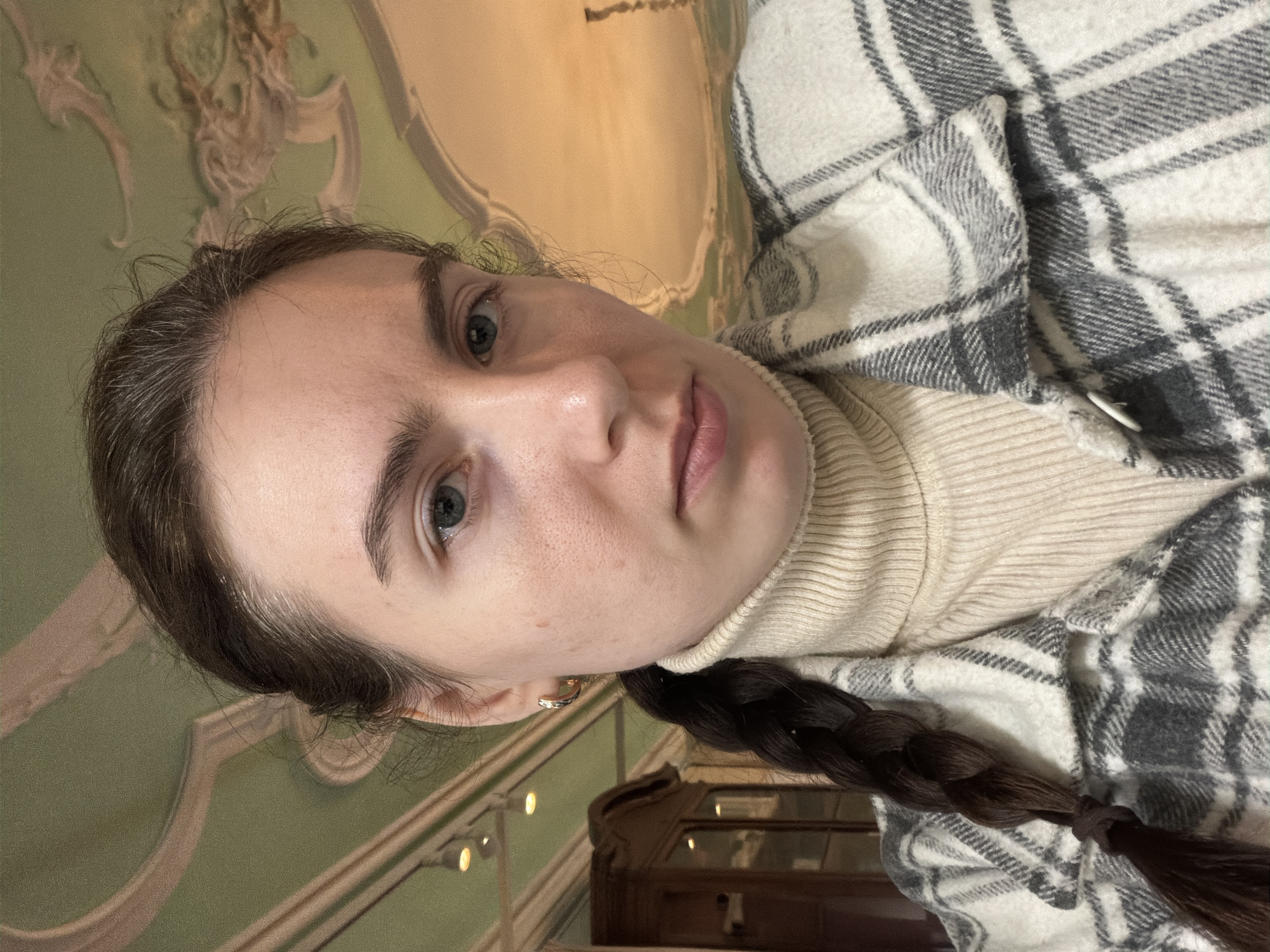 A woman with fair skin, light eyes, and dark brown braided hair is seen in a close-up, slightly angled shot. She wears a cream-colored turtleneck sweater and a grey and white plaid shirt or jacket. She has minimal makeup, and the natural texture of her skin is visible.
The background indicates an interior setting with ornate architectural details. There is light green wall paneling with decorative molding. Hints of a display case or cabinet are visible in the lower part of the frame.
Based on the architectural style, the location is likely a historical building in Saint Petersburg, Russia, known for its elaborate palaces and museums with such interiors.