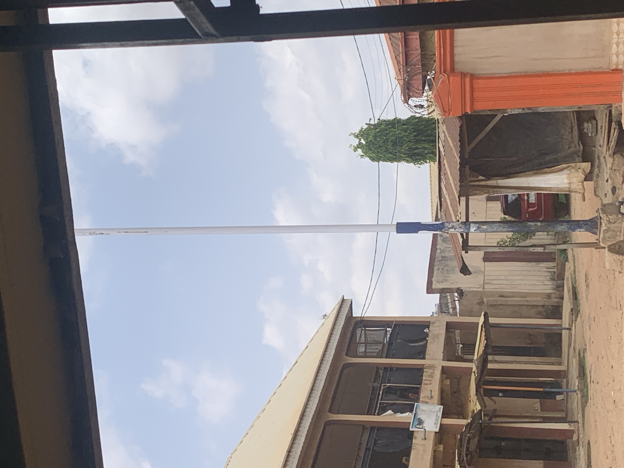 The image shows a street view in Sabon Tasha, Nigeria, partially framed by a dark structure, possibly a building or shelter. A tall white pole extends vertically through the frame, situated along the road. Buildings line the street, including a multi-story structure with open archways and a beige facade. A red vehicle is visible in the background near buildings with orange accents. A tall, thin green tree stands near the buildings, with electrical wires strung across the sky. The sky is light blue with scattered clouds.