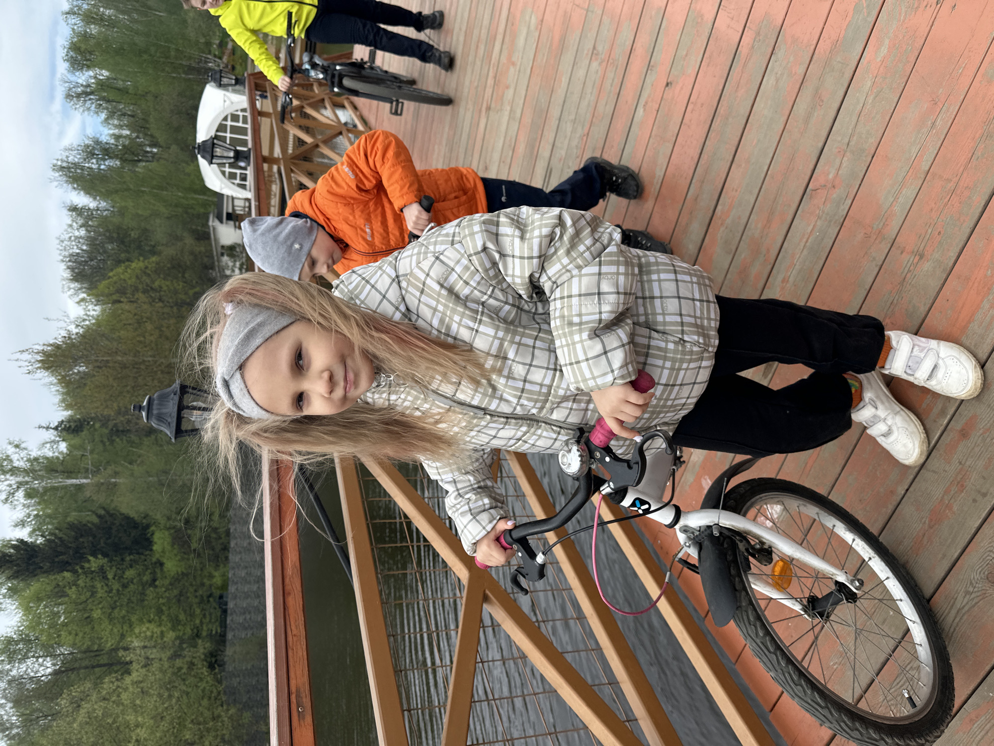 A young girl is standing on a wooden pier, gripping the handlebars of a white bicycle. She wears a plaid jacket, black pants, white sneakers, and a gray headband. Her hair is blonde with some pink streaks. Behind her, another child in an orange jacket lies prone on the pier. Further in the background, a person in a yellow jacket stands near a bicycle. The pier's wooden planks are visible, and a wooden railing separates the pier from the water. Trees are visible across the water, and a white building is partially visible in the distance. The location is Grebnevo, Russia.