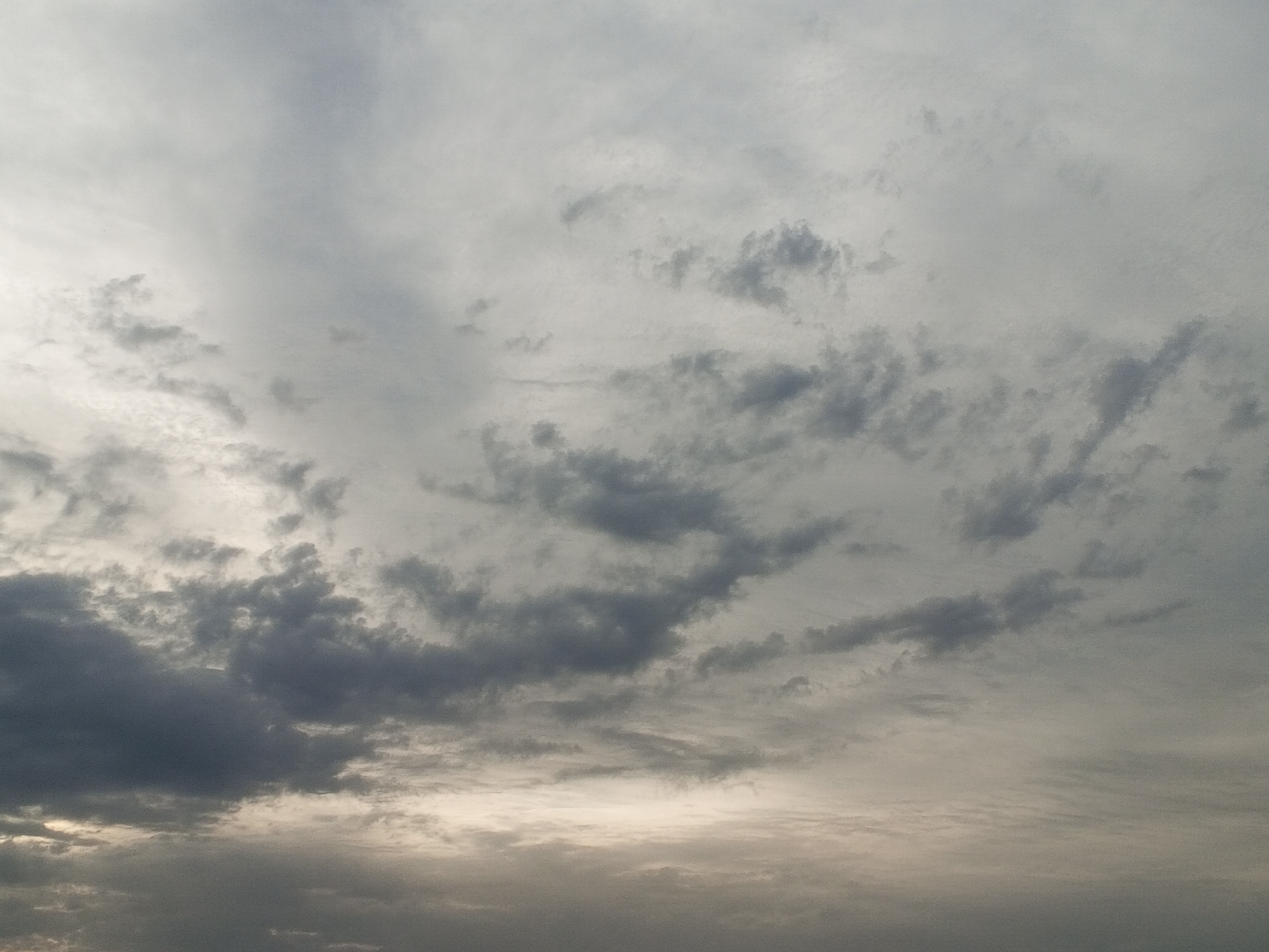 The image captures an expansive view of a cloudy sky, dominated by various shades of grey and white. The scene presents a textured canvas of clouds, ranging from light, wispy formations to denser, darker masses.
Throughout the frame, different types of clouds are visible. Lighter, more diffused clouds form a general overcast layer, particularly noticeable in the upper and right portions of the original image and some of the crops. Interspersed among these are darker, more defined cloud clusters, particularly prominent in the lower-left and central areas of the main image. These darker patches suggest greater thickness or lower altitude.
A notable detail is a brighter, slightly lighter band extending across the bottom-center of the primary image, hinting at the sun's position behind the clouds, possibly indicating early morning or late afternoon light, or simply a break in the cloud cover allowing more light to filter through. The overall lighting is subdued, creating a soft, diffused atmosphere.
There are no people, objects, or visible activities depicted in the media file. No text is present. The weather appears to be overcast or partly cloudy, suggesting a calm or possibly pre-rainy day. While the location context is provided as Badagry, Nigeria, there are no specific geographical cues within the image itself to confirm this.