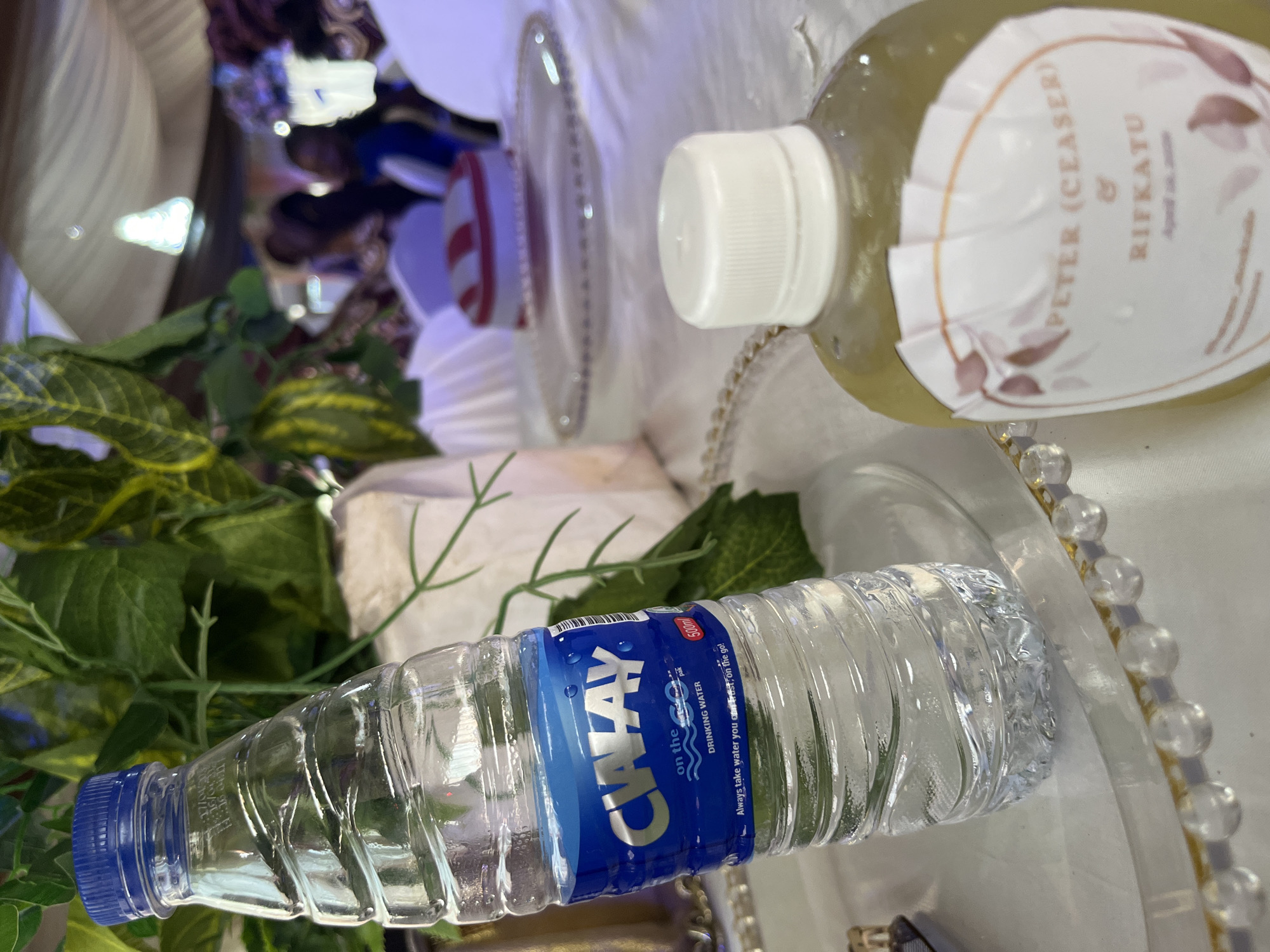 This eye-level shot captures a close-up of a table setting at what appears to be an indoor event or celebration in Abuja, Nigeria. The foreground is dominated by two bottles resting on a white tablecloth, which is adorned with a decorative plate featuring a gold-beaded rim. Artificial green foliage and plant stems are scattered across the table, adding a decorative touch.
One prominent object is a clear plastic bottle of CWAY drinking water, featuring a blue screw-top cap. The blue label on the bottle is clearly visible and reads "CWAY on the go! DRINKING WATER" and "Always take water you can trust on the go!" It also indicates "500ml".
Next to the CWAY bottle is another clear bottle, this one containing a pale yellow liquid, possibly a custom drink or juice. It has a white screw-top cap and a personalized label. The legible text on this label identifies "PETER (CEASER) & RIFKATU," suggesting it's a memento or customized item for a wedding, engagement, or similar special occasion involving these two individuals.
The background is softly blurred, showing glimpses of other table settings, white fabric draping, and indistinct shapes that suggest people and other event decor, reinforcing the setting as a festive gathering. The lighting is bright and even, consistent with an indoor event. The presence of the CWAY brand, a well-known Nigerian company, aligns with the provided location context of Abuja, Nigeria.