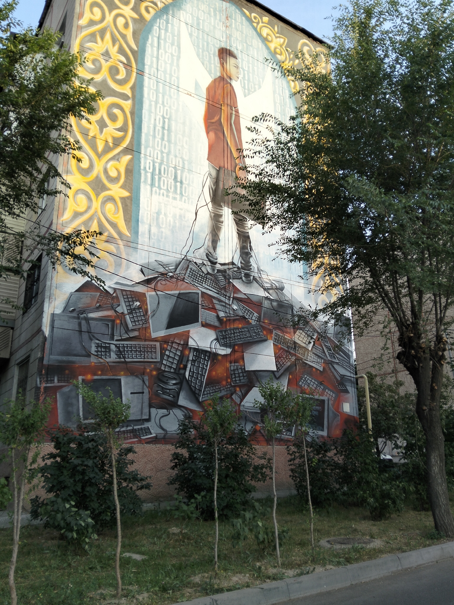 A mural adorns the side of a multi-story building in Almaty, Kazakhstan. The mural features a stylized depiction of a young man standing atop a pile of computer monitors and keyboards. The background of the mural incorporates elements of traditional Kazakh ornamentation, such as swirling yellow patterns, juxtaposed with binary code and an abstract archway. The building itself appears to be constructed of concrete and is partially obscured by trees and power lines. The ground level shows a grassy area with small saplings and bushes, bordering a paved road. The sky is visible in the upper portion of the image.