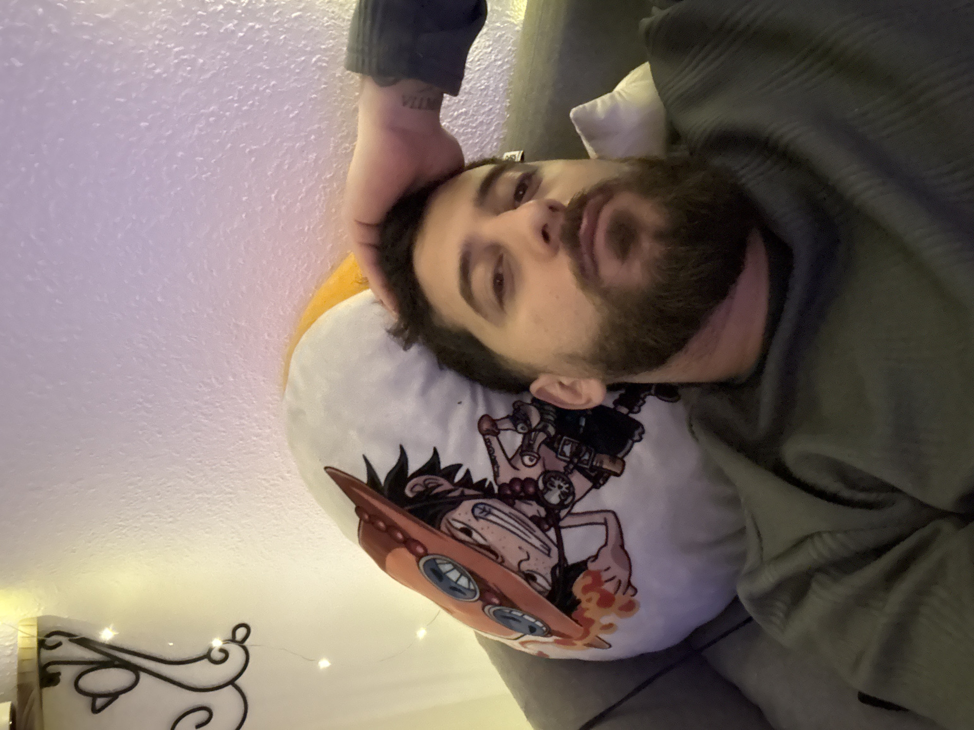 An adult male is depicted from the chest up, positioned lying back against a dark gray upholstered surface, likely a sofa or chair. He has light skin, dark hair, and a full dark beard and mustache. His left hand rests on the top of his head, with a faint tattoo visible on his left forearm. He is wearing a dark green, ribbed, long-sleeved garment and appears to be looking directly at the camera with a neutral expression. Behind his head is a white, round cushion featuring a colorful cartoon illustration of Portgas D. Ace from One Piece, identifiable by his distinctive orange hat with skull-like buttons, dark spiky hair, freckles, and beaded necklace, depicted with fiery effects around his hand. The background consists of a light-colored, textured wall. Warm string lights with small points of light are visible in the upper left corner, attached near a dark, ornate metal wall decoration or shelf bracket. A cooler, purplish light illuminates parts of the upper right wall. The image does not contain any discernible visual information to identify its location as Strasbourg, France.