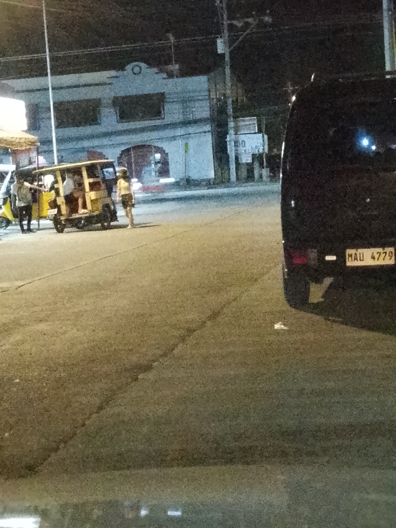 The image depicts a nighttime scene on a street in Davao City, Philippines. The primary subjects are people and vehicles. In the foreground, a dark-colored van is parked, partially obscuring the view of a yellow tricycle. The van has a license plate that reads "MAU 4779". The tricycle is carrying several passengers and is illuminated by external lights. A woman in a yellow top and dark shorts stands near the tricycle, appearing to be interacting with someone or observing the scene. Another person, dressed in black and white, stands to the left of the tricycle, possibly a driver or passenger.
In the background, there's a building with a distinct architectural style, featuring what appears to be arched windows or doorways. Signs are visible on a pole near the building, though the text is partially obscured and grainy. The street is paved, and the overall lighting suggests it is nighttime. The presence of the tricycle, a common mode of public transportation in the Philippines, and the general atmosphere of the scene are indicative of a street in a Philippine city. There are no clear indicators of the weather, but the darkness suggests it is not daytime. The scene appears to be a mundane street encounter, possibly involving the loading or unloading of passengers.