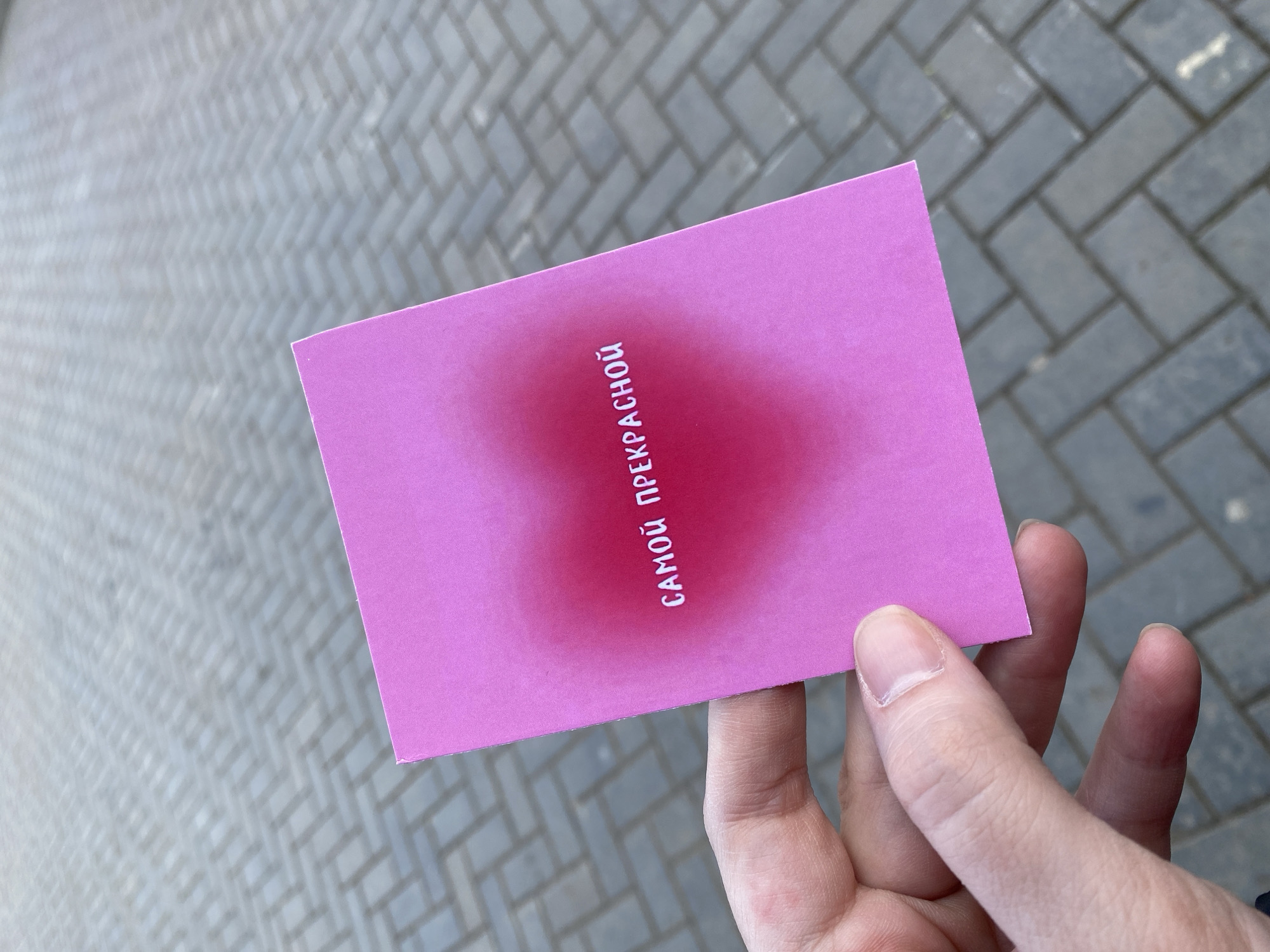 A hand holds a pink card with the text "САМОЙ ПРЕКРАСНОЙ" printed on it vertically. The card has a darker pink gradient in the center. The background consists of a gray brick walkway. While the exact location cannot be determined from the image alone, the image has a geotag identifying Votkinsk, Russia as the location.