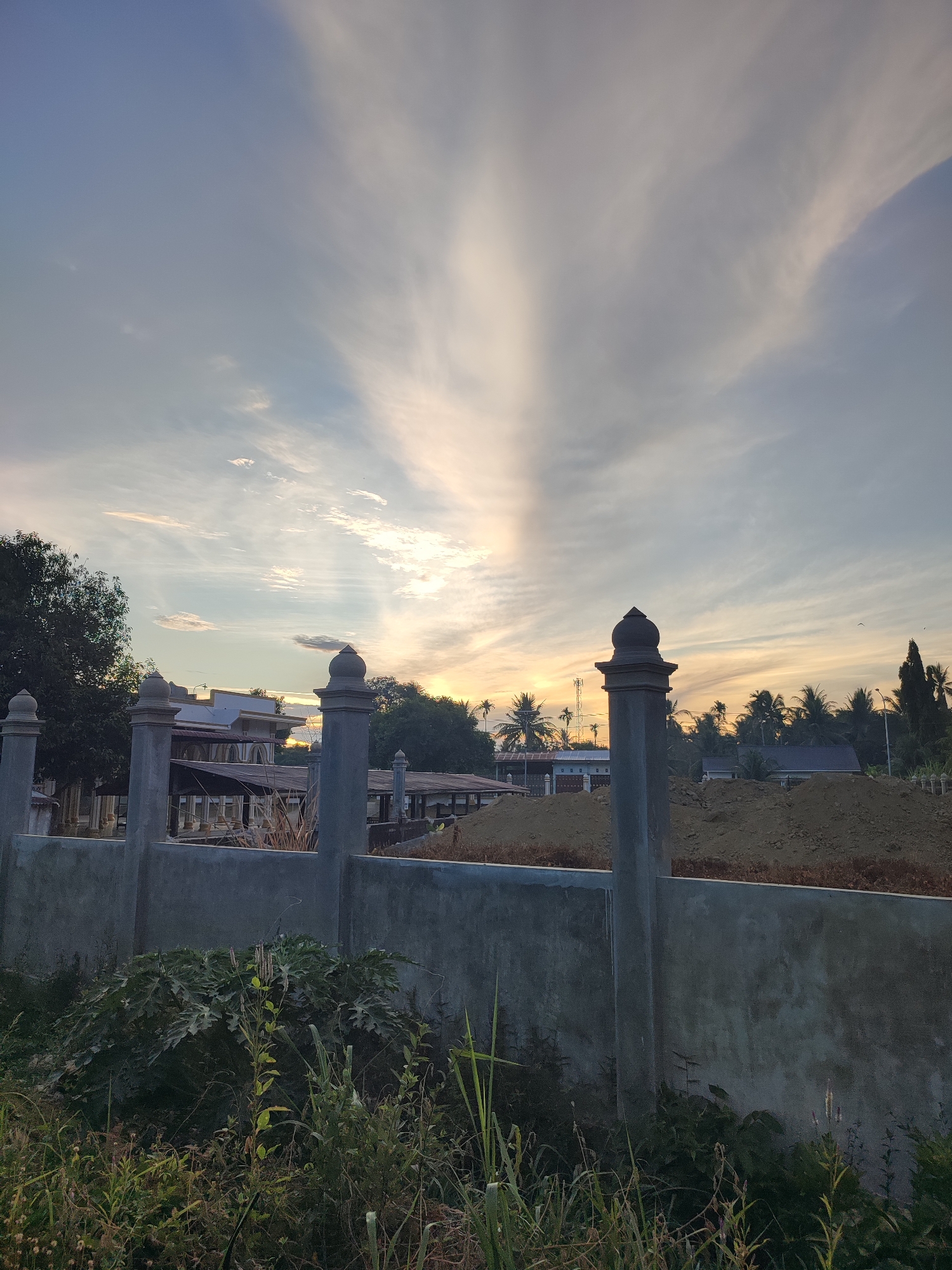 The image captures an outdoor scene at dawn or dusk, likely in Gampongrayadagang, Indonesia, characterized by a serene sky with elongated clouds radiating from a central point, suggesting the sun is near the horizon. In the foreground, a low, weathered concrete wall with decorative pillar caps runs horizontally across the frame. Beyond the wall, vegetation is visible, including tall grasses and leafy plants. Further back, a cluster of buildings with sloped roofs and intricate architectural details are partially obscured by trees. The landscape features palm trees and other tropical foliage, indicative of a warm climate. A large pile of earth or construction material is also present. The overall impression is of a quiet, possibly rural or semi-rural setting during the transitional hours of the day. There are no people or animals visible in the image.