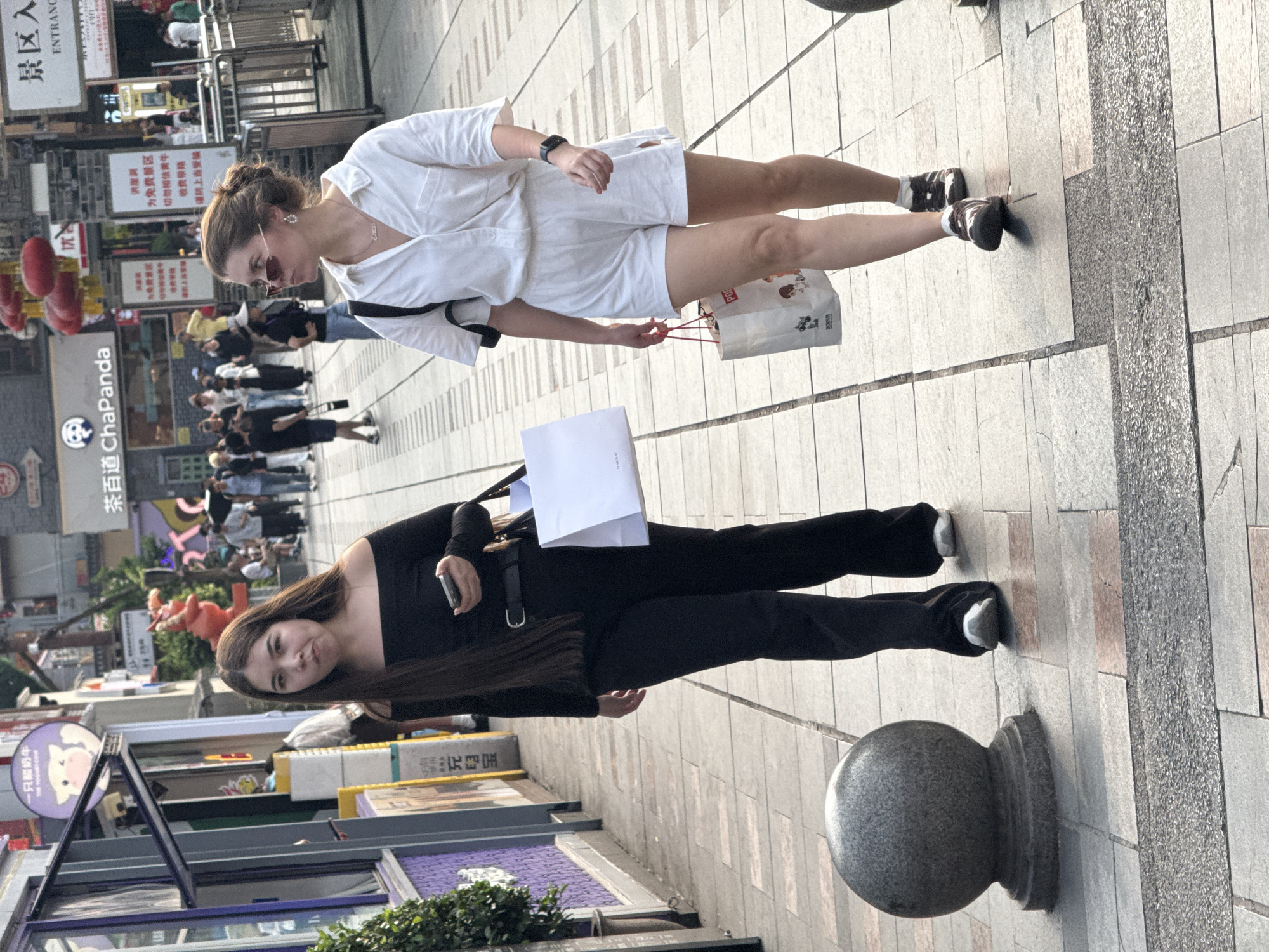 A street scene in Yuzhong District, China, depicts two women standing on a pedestrian walkway. The woman on the left is wearing a black long-sleeve top and black pants, and she holds a white paper bag in her left hand while holding a smartphone in her right hand. The other woman is wearing a white button-down shirt and white shorts; she is carrying a shopping bag in her right hand. The pedestrian walkway is composed of tiled pavement. In the background, buildings and storefronts can be seen, including a storefront with a panda logo and another with Chinese characters. A decorative stone ball is on the bottom right of the frame.