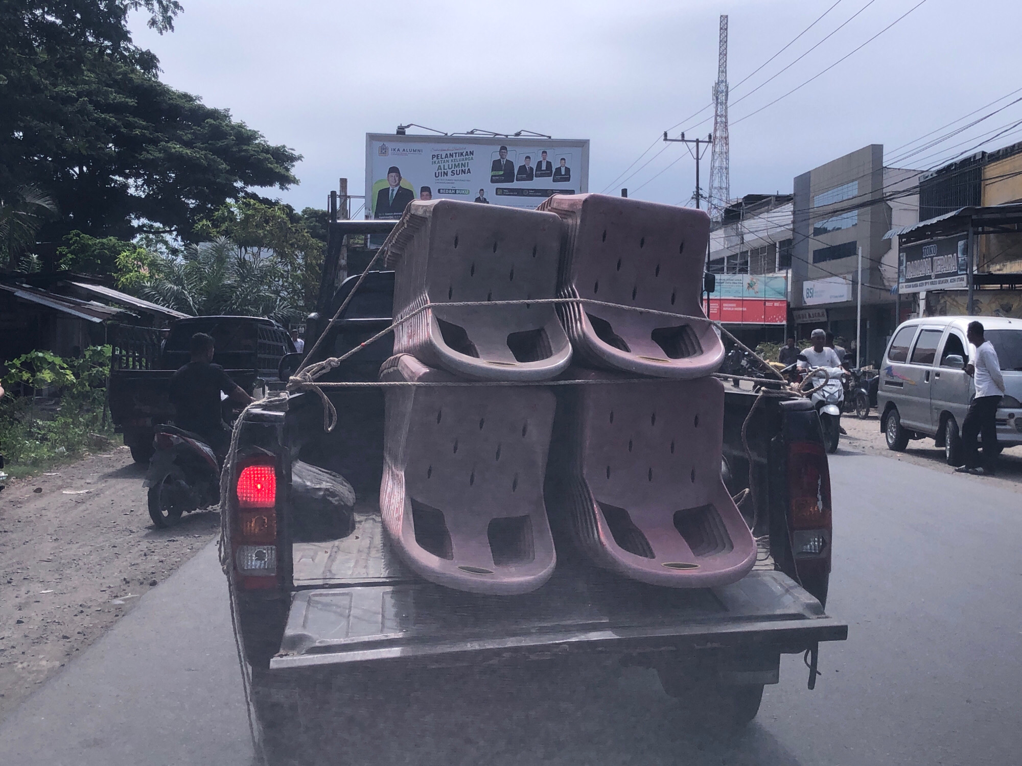 A pickup truck is transporting several large, reddish-pink plastic seats in its bed. The seats are stacked and secured with ropes. The truck is on a paved road, with other vehicles visible in the background, including a silver van, a motorcycle, and another dark-colored truck. Buildings and a large billboard are also present along the roadside. The environment appears to be urban, with overhead power lines and a telecommunications tower in the distance. The sky is overcast. The scene is consistent with a street in Palohlada, Indonesia, given the presence of typical urban infrastructure and vehicles found in the region.