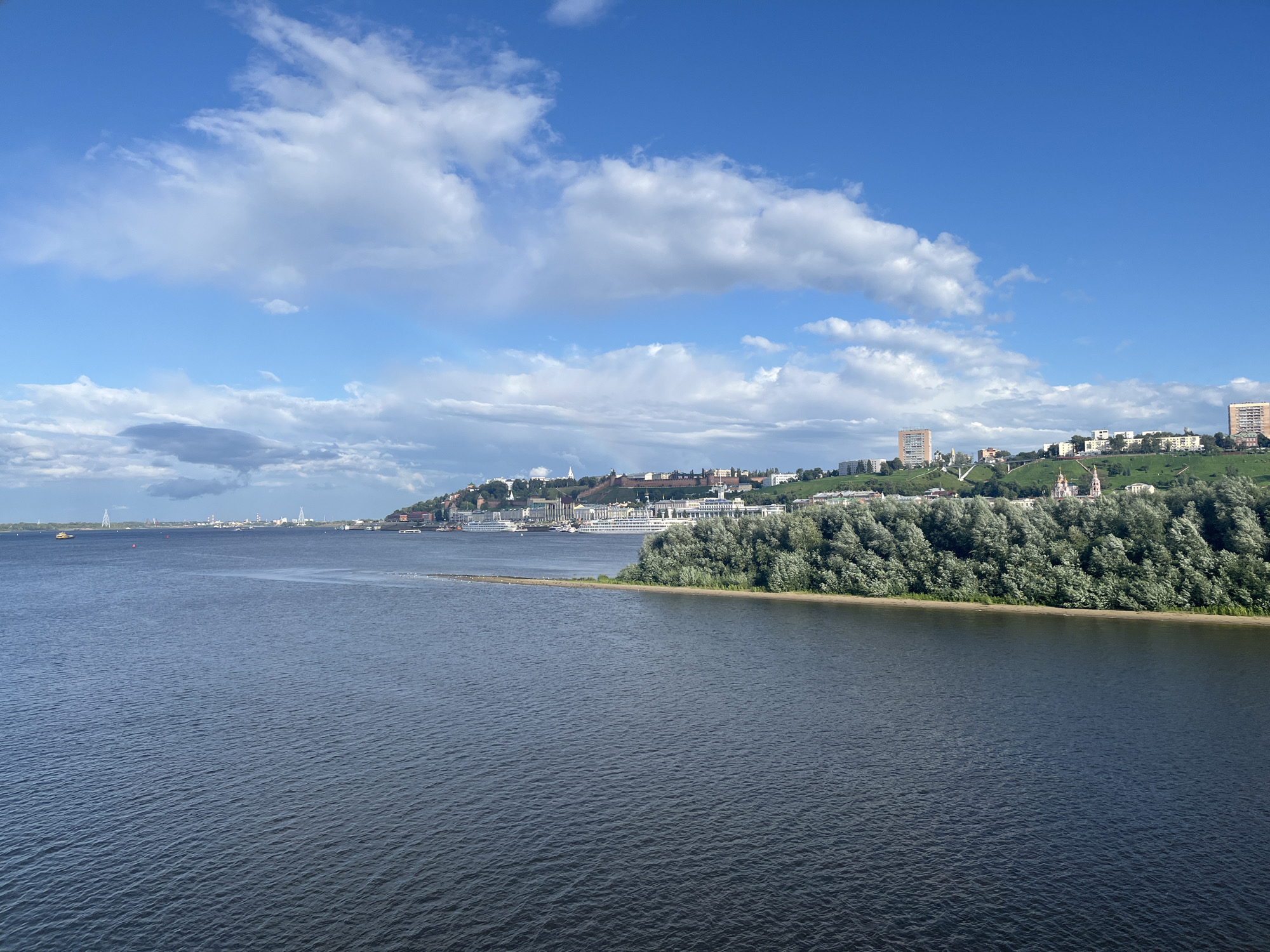 This is an eye-level, wide shot of the Volga River in Nizhny Novgorod, Russia, on a partly cloudy day. The river takes up the lower two-thirds of the frame, with choppy, dark blue water. In the distance, on the far bank, a city skyline rises above a lush, green hillside dotted with trees. The city features numerous buildings of various sizes and architectural styles, including tall, modern apartment blocks and older, more ornate structures. Several cruise ships are docked along the riverfront, their white hulls stark against the water. The sky is a vibrant blue, with scattered white, fluffy clouds. A sandy spit of land, partially submerged in the river, juts out from the right side of the frame, covered with dense green foliage. There are no people visible in the image.