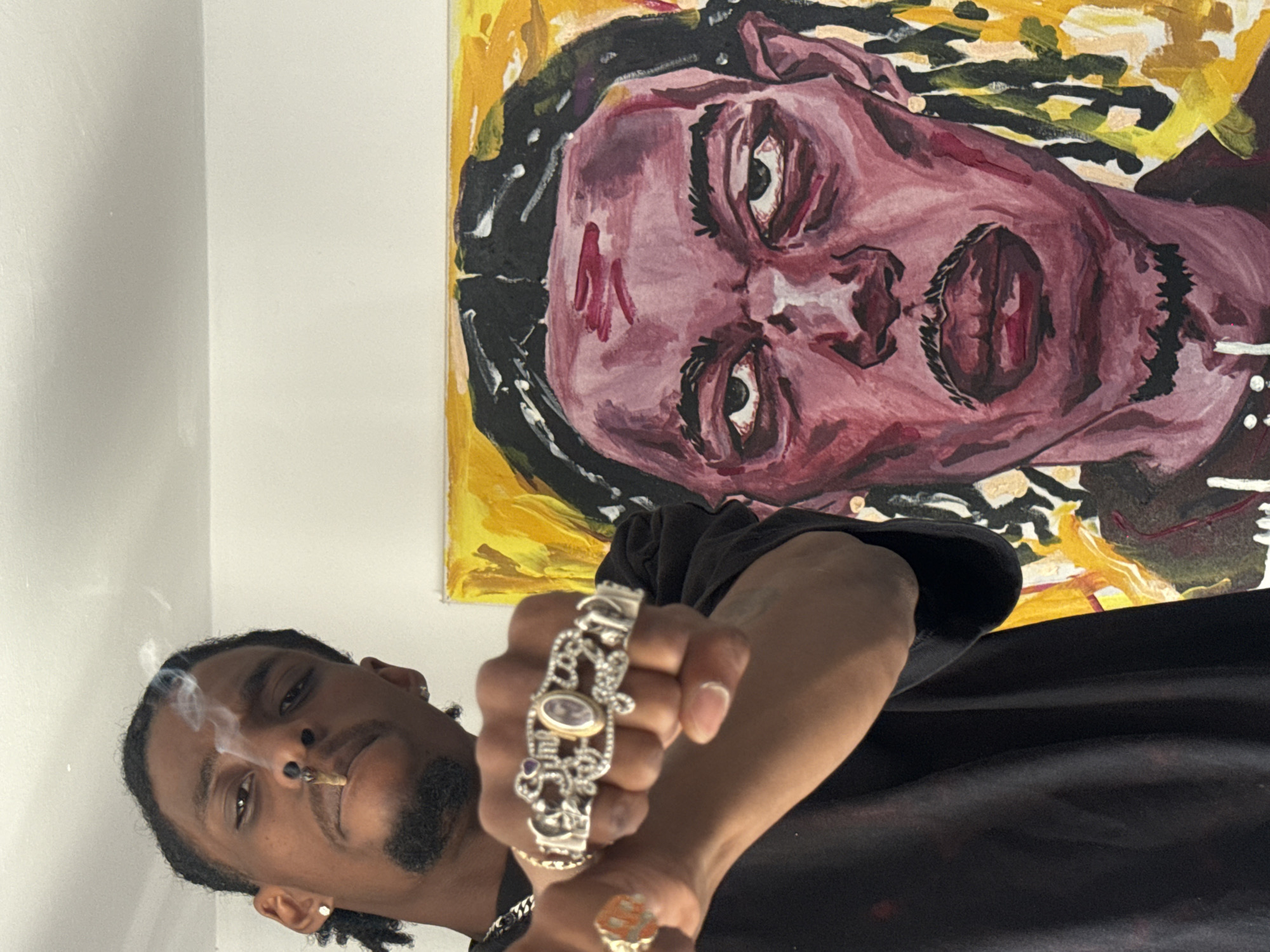 A portrait-style photograph, rotated clockwise, captures a young man posing indoors against a plain white wall, likely in London, United Kingdom. In the foreground, the man, depicted from a low angle, has dark skin, dreadlocks or braids, a mustache, and a goatee beard. He wears a nose ring in his left nostril and a stud earring in his left earlobe. A lit cigarette or joint is in his mouth, with smoke gently rising around his face. His right arm is extended towards the viewer, showcasing an ornate, silver-colored bracelet with oval stones and intricate metalwork, along with several rings on his fingers, including a prominent patterned one on his index finger and a gold-toned ring on his pinky. He is dressed in a dark, possibly black, long-sleeved shirt and looks directly at the camera with a calm, focused expression.
In the background, a vibrant, expressive portrait painting hangs on the white wall. The artwork features a stylized face with striking eyes, rendered predominantly in shades of maroon and red for the skin, with bold black outlines and hair. The background of the painting is a vivid yellow or gold, with abstract touches of green and white brushstrokes, contributing to its raw, artistic style. The indoor setting, characterized by the simple white walls, provides a minimalist backdrop that highlights both the subject and the artwork. The time of day is not discernible, and there is no visible text in the image.