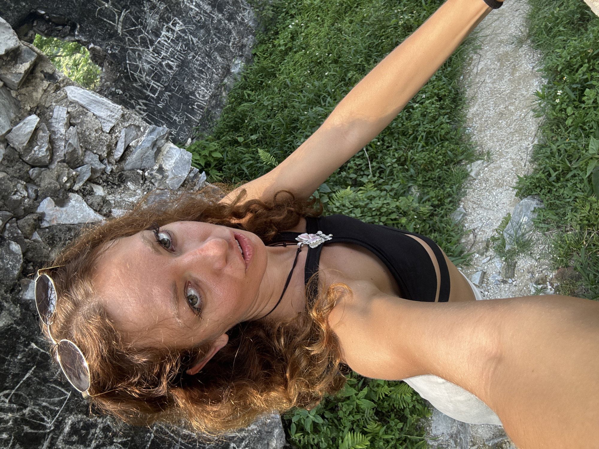 A woman with curly auburn hair wearing a black top and sunglasses stands near a stone structure with graffiti. Green foliage surrounds the structure and extends to a path. The location is Đường Thượng Commune, Vietnam.