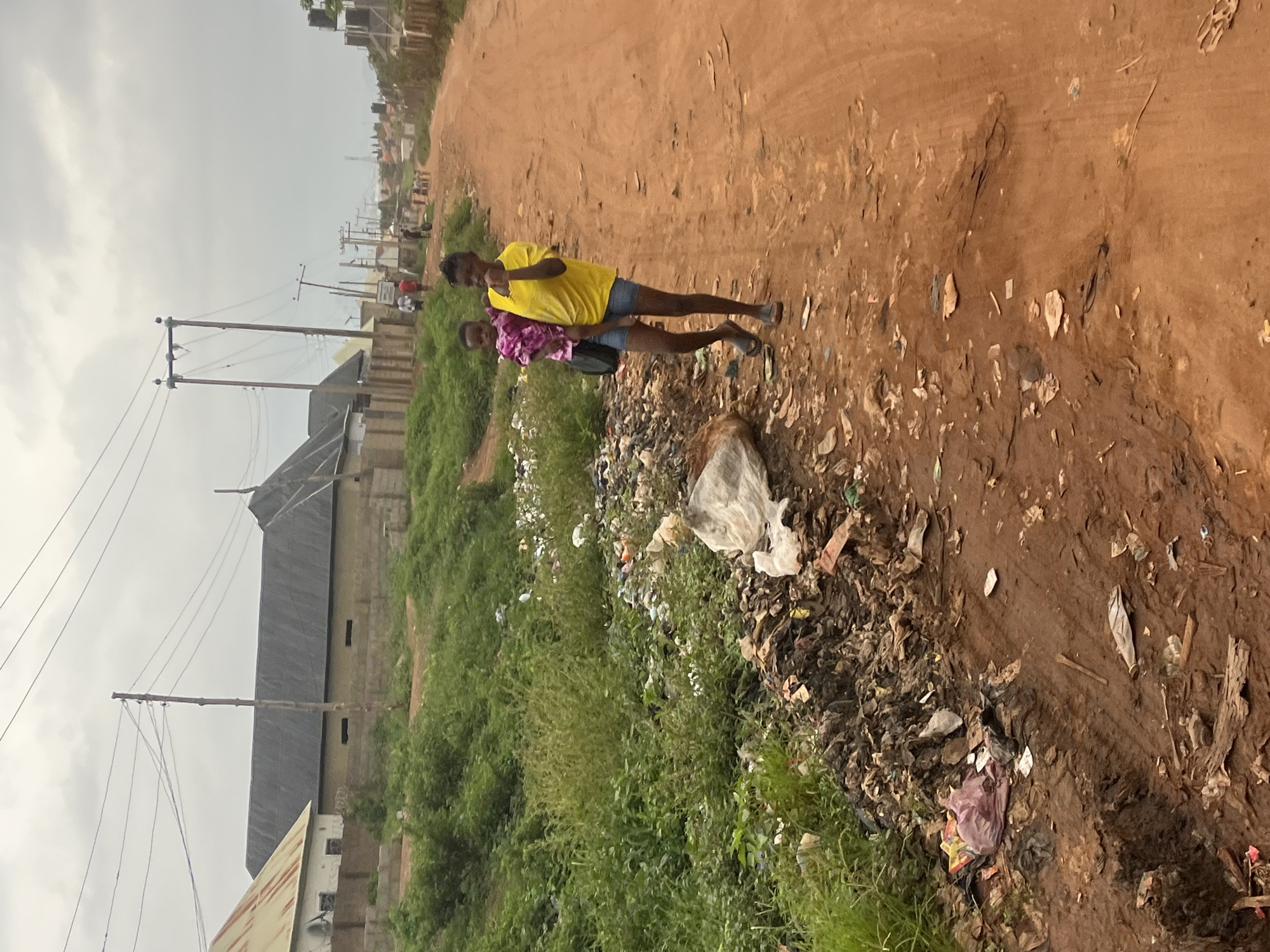 The image, taken in Abuja, Nigeria, captures an outdoor scene dominated by an unpaved, reddish-brown dirt road under an overcast daytime sky. Two young individuals are central subjects, standing on the road. The person in front wears a bright yellow t-shirt and blue denim shorts, while the one slightly behind them is dressed in a pink and white patterned top and blue denim shorts, carrying a dark shoulder bag. Both appear to be looking forward, possibly towards the camera, with neutral expressions.
The scene reveals a significant issue with waste management, as a large amount of litter and debris, including plastic bags and wrappers, is visible along the right edge of the road, spilling into a ditch-like area. On the left side of the road, lush green vegetation, consisting of grass and bushes, borders the path.
In the mid-ground and background, utility poles with numerous overhead power lines are evident, suggesting existing infrastructure. Beyond these, a cluster of residential or commercial buildings can be seen, characterized by simple, block-like construction and dark-colored corrugated roofs. Some of these structures feature elevated water tanks. The overall setting depicts a developing or semi-urban residential area in Abuja, Nigeria. There is no visible text in the image.
