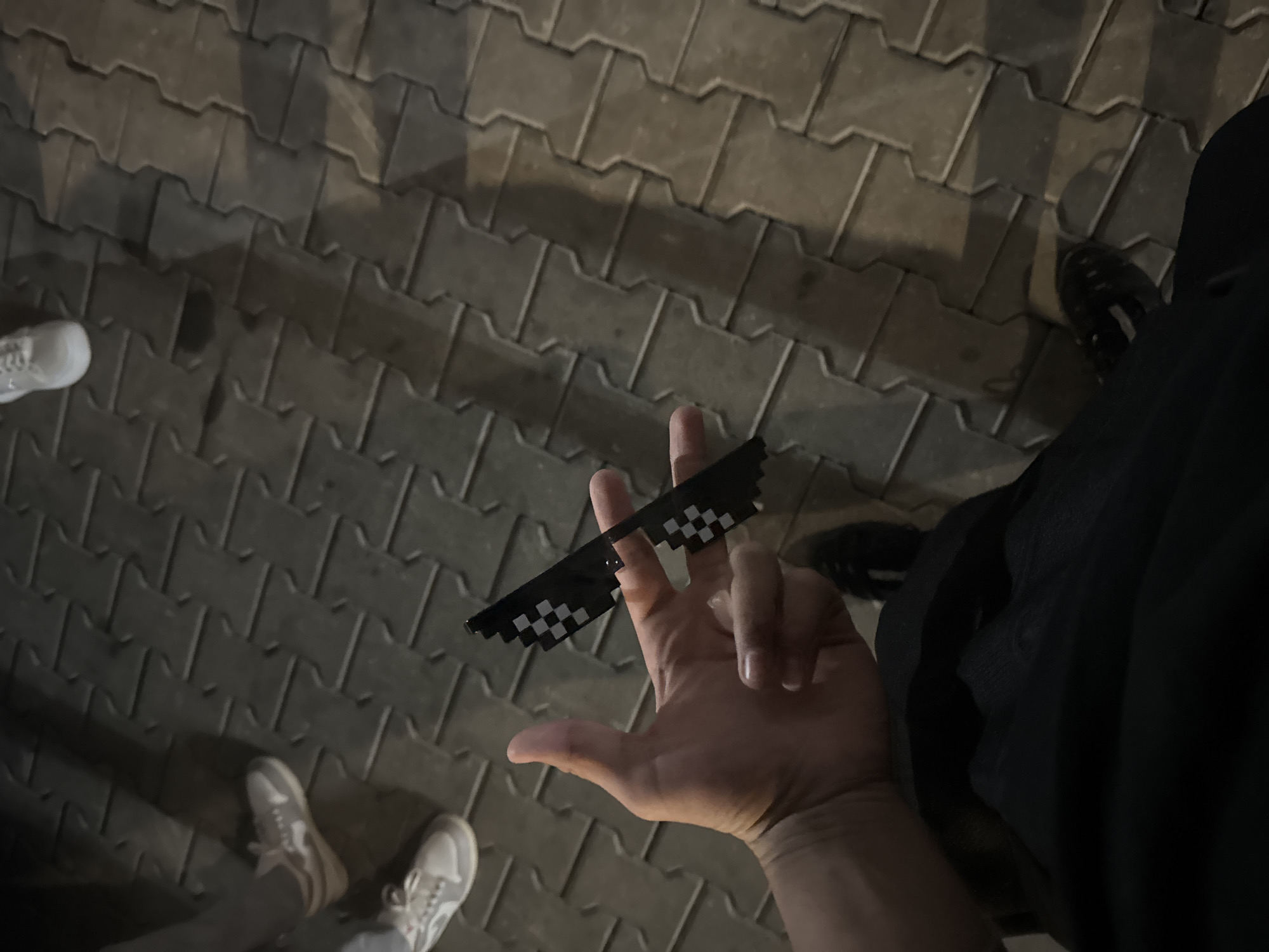 The image captures an outdoor scene at night or in very low light conditions, set on a pavement made of dark grey interlocking paving stones. The main subject is a human hand, illuminated by a light source, holding a pair of black pixelated sunglasses, often associated with the "deal with it" internet meme. The hand is holding the glasses by the top edge with the index finger and thumb.
In the background, the feet of at least two individuals are visible on the pavement. To the left, a pair of light grey or white sneakers is partially shown. To the right, a dark-colored shoe, possibly a boot or dark sneaker, is visible along with a dark pant leg, likely belonging to the person holding the glasses. Long, indistinct shadows are cast across the pavement, consistent with the low light environment.
The scene suggests a casual, perhaps humorous, moment taking place in a public pedestrian area in Sevlievo, Bulgaria. The pixelated sunglasses imply a playful or meme-related activity. No specific weather conditions are discernible, but the pavement appears dry. There is no visible text in the image.