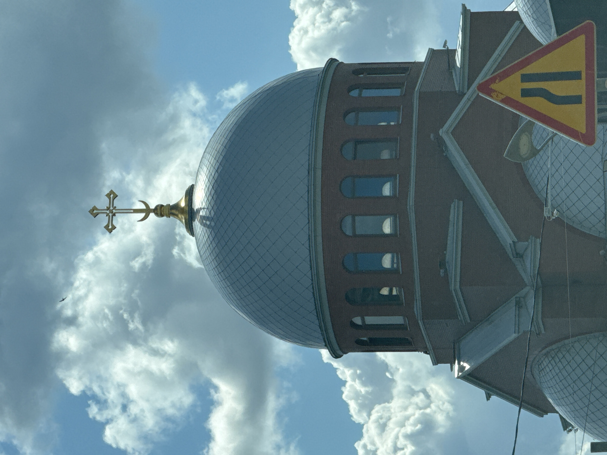 A bright, cloudy sky with scattered white cumulus clouds forms the backdrop for a brick church with a prominent silver dome topped with a gold cross. The church has a series of arched windows encircling the upper portion of its structure. In the foreground, a yellow triangular traffic sign with a red border displays two dark vertical lines, indicating a narrowing of the road ahead. The sign is partially obscuring the lower portion of the church and the sky. A single bird is visible flying in the upper left quadrant of the image against the clouds. The image appears to be taken from a low-angle perspective, possibly from a vehicle, given the presence of the traffic sign and the angle of the shot. The location is identified as Sinyavino, Russia.