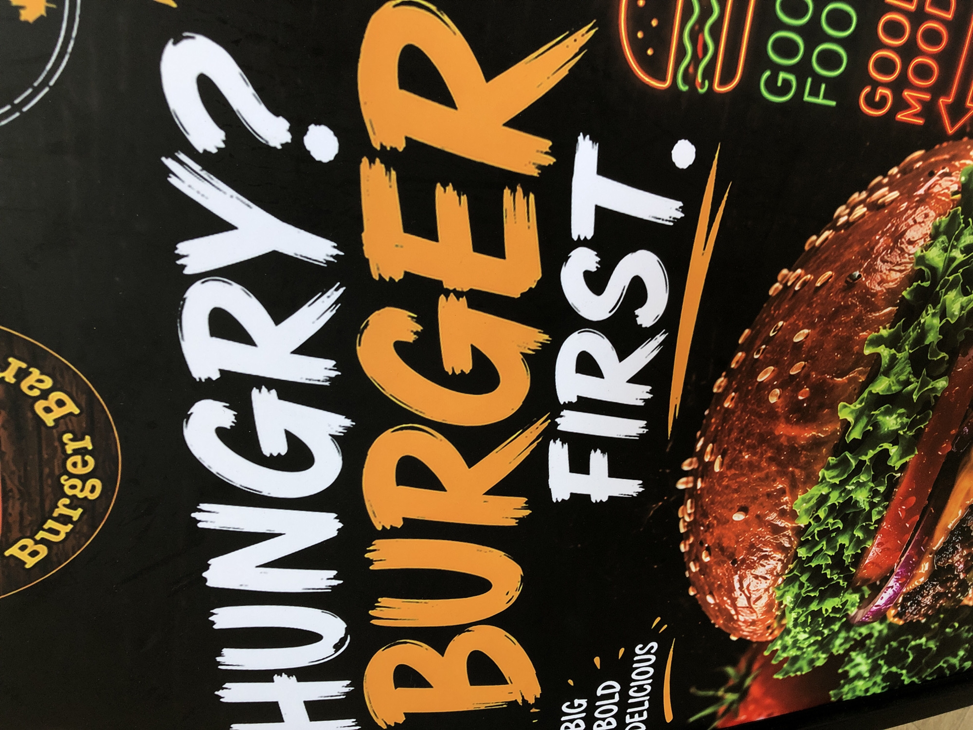 The media file is a close-up shot of a black poster advertising a burger bar. The poster has a bold, graphic design with large, brushstroke-style text and an image of a burger.
The main text on the poster reads "HUNGRY? BURGER FIRST." with "HUNGRY?" in white and "BURGER FIRST." in orange. There are also smaller text elements. To the left, in a circular emblem, the words "Burger Bar" are visible in yellow text. To the right, there are neon-style graphics, including a stylized burger in green and red, and text that reads "GOOD FOOD GOOD MOOD" in a similar neon style, with "GOOD" in green and "MOOD" in orange. Below the main text, there is more small text that appears to read "BIG BOLD DELICIOUS" in white.
The background is solid black, which makes the vibrant text and the image of the burger stand out. The burger itself is a prominent feature, showing a close-up of its components: a dark bun covered in sesame seeds, fresh green lettuce, and red tomato slices. The overall impression is one of a lively and appetizing advertisement for a burger restaurant. The style suggests a modern and casual dining experience. The location context of Da Nang, Vietnam, is not directly evident from the image itself but is provided as external information. There are no people or specific weather conditions depicted. The image focuses solely on the promotional material.