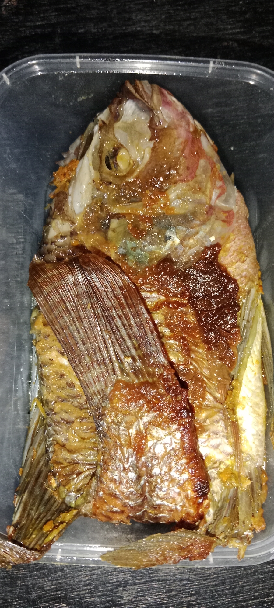 A close-up, overhead shot shows a whole, cooked fish, likely a type of tilapia, presented in a clear plastic container. The fish is golden brown and appears to be seasoned with a dark spice mixture, visible as reddish-brown patches across its body and around the head. The fish has been split lengthwise and is lying on its side, revealing its internal structure. The eye of the fish is visible, along with the gill area. The tail fin is partially obscured by the body. The fish is resting on a dark, textured surface, possibly a wooden table, with the edge of the plastic container visible in the foreground and background. The image is well-lit, highlighting the glistening texture of the cooked fish. There are no people or any indications of location within Subang, Indonesia, present in the image.
