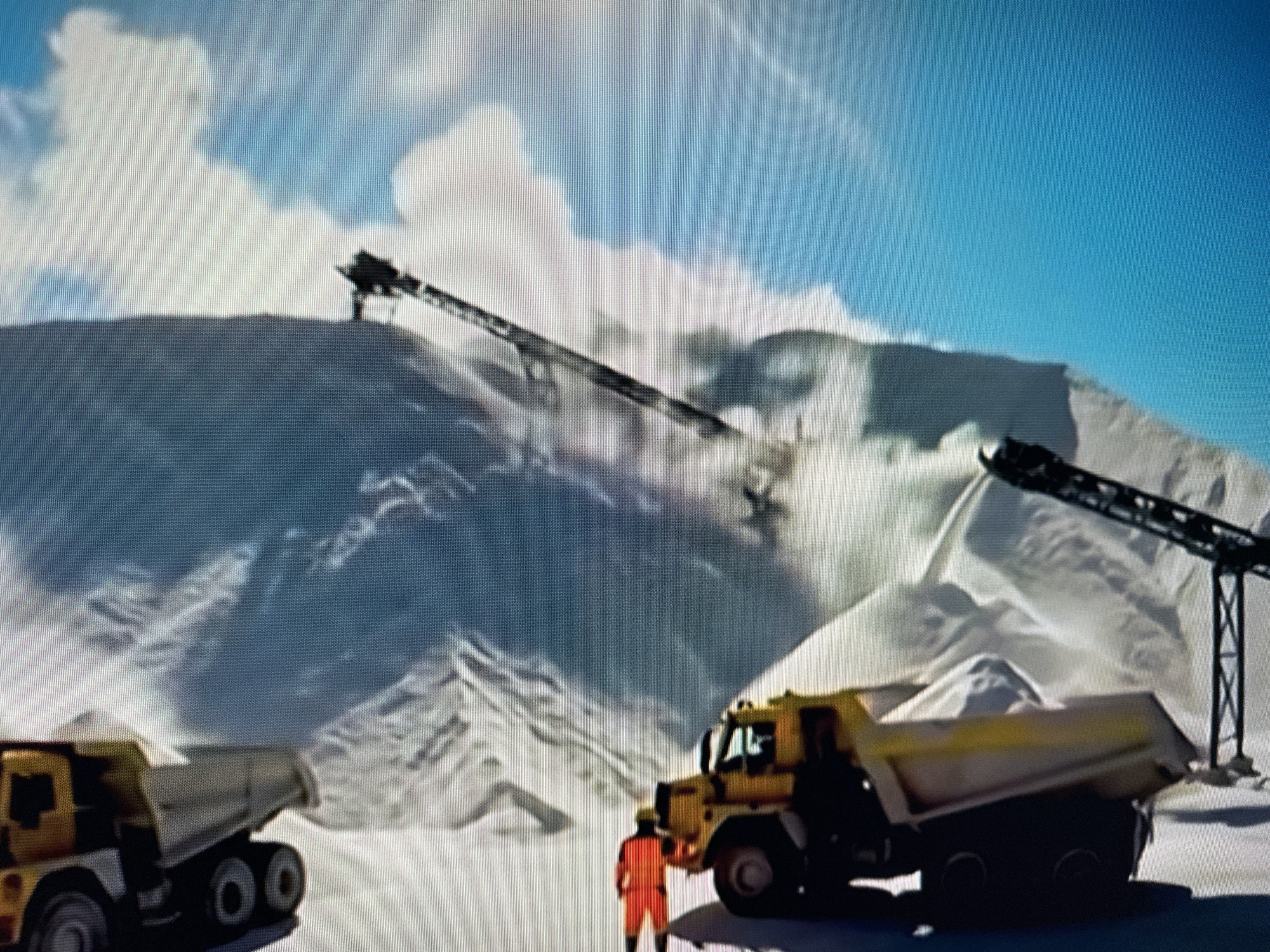 This is a digitally rendered image of a mountainous, snow-covered landscape under a bright blue sky with scattered white clouds. In the foreground, two yellow dump trucks are parked. One truck is partially visible on the left, and a larger yellow dump truck with a load of white material is on the right, facing left. A person wearing an orange jumpsuit is standing between the two trucks, facing the larger truck. In the background, two conveyor belts extend from the upper right and upper left portions of the image, seemingly depositing material onto the mountain slopes. The environment suggests an industrial or mining operation in a cold, mountainous region. The image does not depict any recognizable landmarks or features of the city of Krasnodar, Russia, nor is there any information to suggest it is located there.