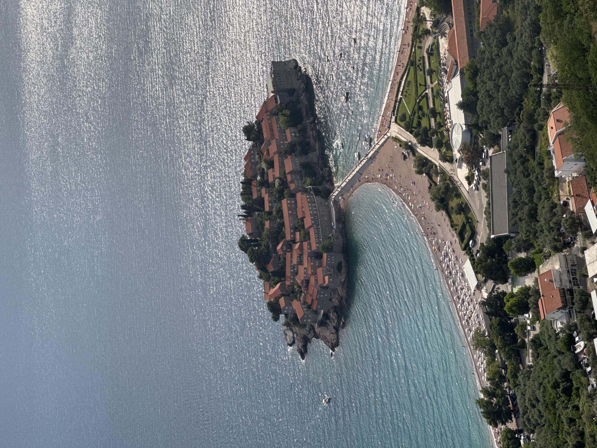An aerial, high-angle shot captures the island town of Sveti Stefan, Montenegro, connected to the mainland by a narrow causeway. The island is densely packed with red-roofed buildings and is surrounded by the shimmering, blue Adriatic Sea. A sandy beach, dotted with numerous white umbrellas and people, curves from the mainland towards the island. Beyond the beach, lush green trees and gardens are visible, interspersed with buildings and parking lots. The water exhibits a texture of small waves, with sunlight reflecting off its surface. Several small boats are present in the water. The scene is bathed in bright sunlight, casting shadows and highlighting the vibrant colors of the landscape.