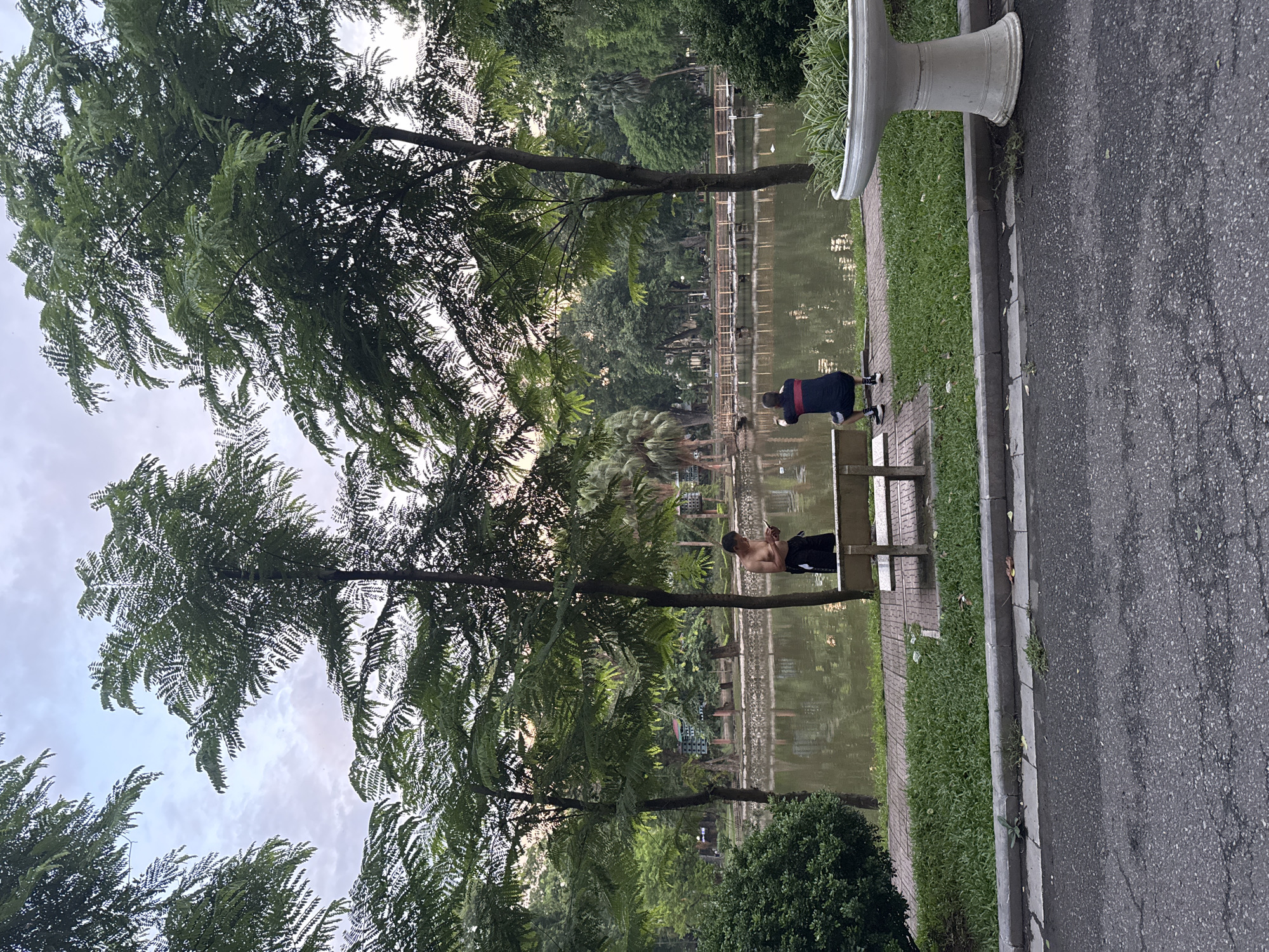 A medium shot captures a serene park scene in Hanoi, Vietnam, near a body of water. In the foreground, the asphalt of a pathway is cracked and weathered. Beyond it, a strip of green grass meets a patterned brick walkway. A white planter with lush green foliage sits to the right. A wooden bench is positioned on the brick walkway, facing the water.
Two individuals are present. A shirtless man, wearing black shorts, stands to the left of the bench, looking down at his phone. To the right of the bench, a person in a dark blue shirt with red and white stripes on the shoulders and black shorts crouches, appearing to take a photo with a mobile device.
The park features several tall trees with dense green foliage, interspersed with smaller palm trees and other lush vegetation. In the background, a calm body of water reflects the trees and the sky. A wooden bridge with a railing spans across the water in the mid-ground. The sky is overcast with hints of light peeking through the clouds. The overall atmosphere suggests a quiet, early morning or late afternoon in a city park.