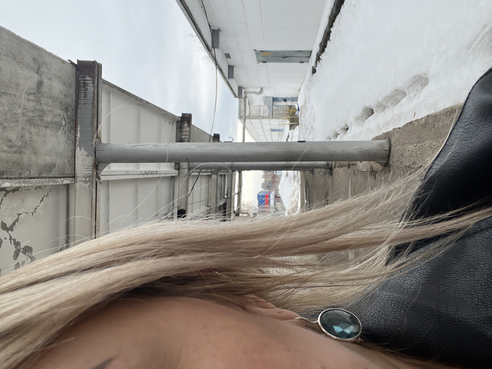The image depicts a partial, close-up view of an individual's left side in the foreground, set against an industrial or utilitarian outdoor environment in Chaikovsky, Russia, during what appears to be winter or early spring. The individual has blonde hair, fair skin, and a dark eyebrow, wearing a black leather-textured garment. A silver-framed, dark teal, oval-shaped earring is visible on the left earlobe.
The background features a concrete-covered walkway supported by multiple grey cylindrical pillars that recede into the distance on the left. The concrete roof and support beams show weathering, discoloration, and some rust. Alongside the walkway, a narrow path runs adjacent to a snow-covered ground with visible footprints and remnants of partially melted snow.
To the right, a series of white, single-story industrial-style buildings are visible. The closest building has a light grey pitched roof, white exterior walls, and a light grey double door. Further back, another white building with blue doors is partially obscured. Beyond the structures, sparse, bare trees are visible under an overcast sky. In the far distance, a red, white, and blue colored structure is faintly discernible.
