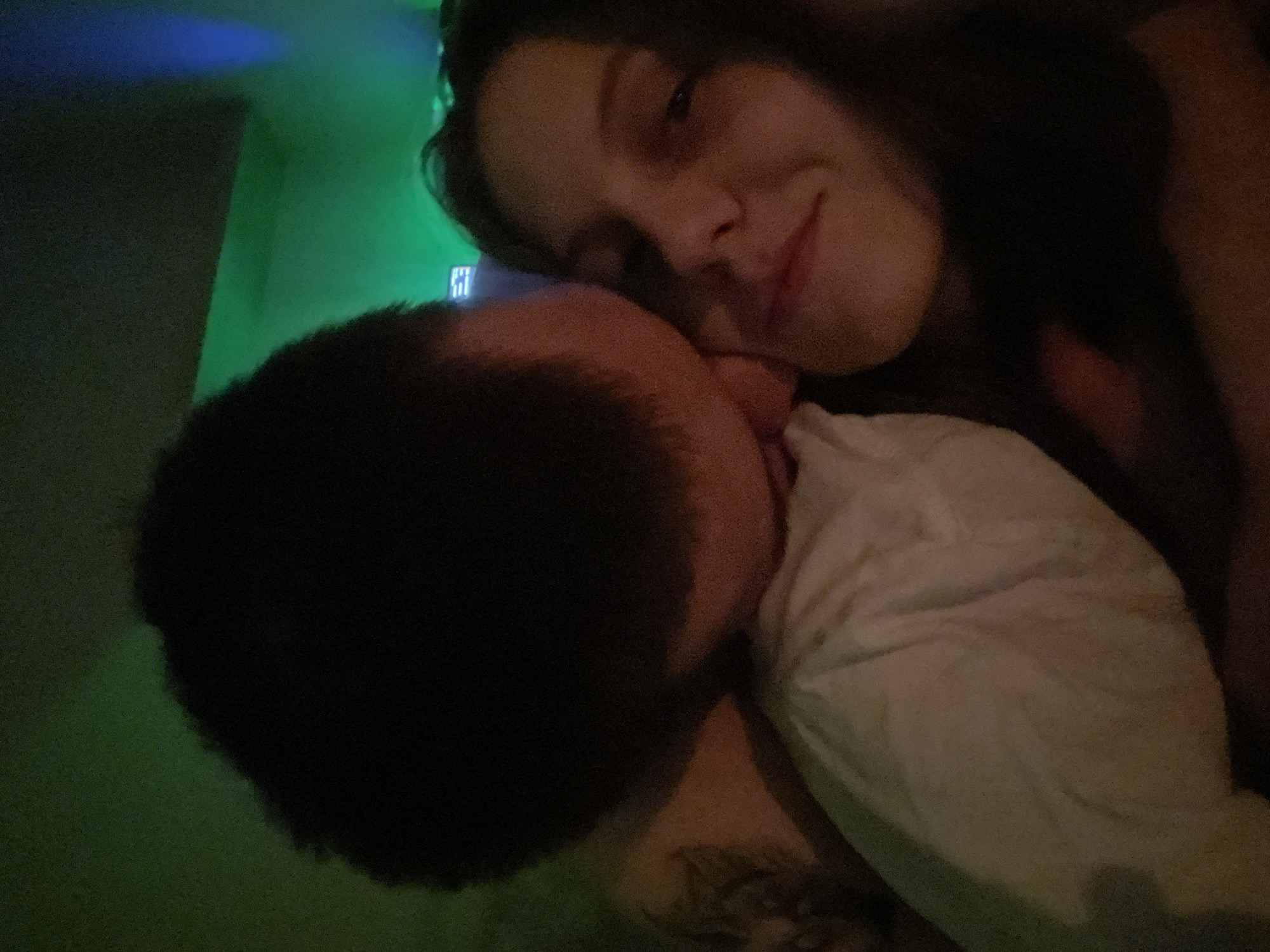 A woman with long brown hair is embracing a person with short, dark hair. The woman is looking at the camera with a slight smile. The other person is wearing a white shirt. A tattoo is partially visible on the shoulder of the person wearing the white shirt. The background is green with a blue light in the upper left corner. The location in the city of Ufa, Russia is not discernible from this image.