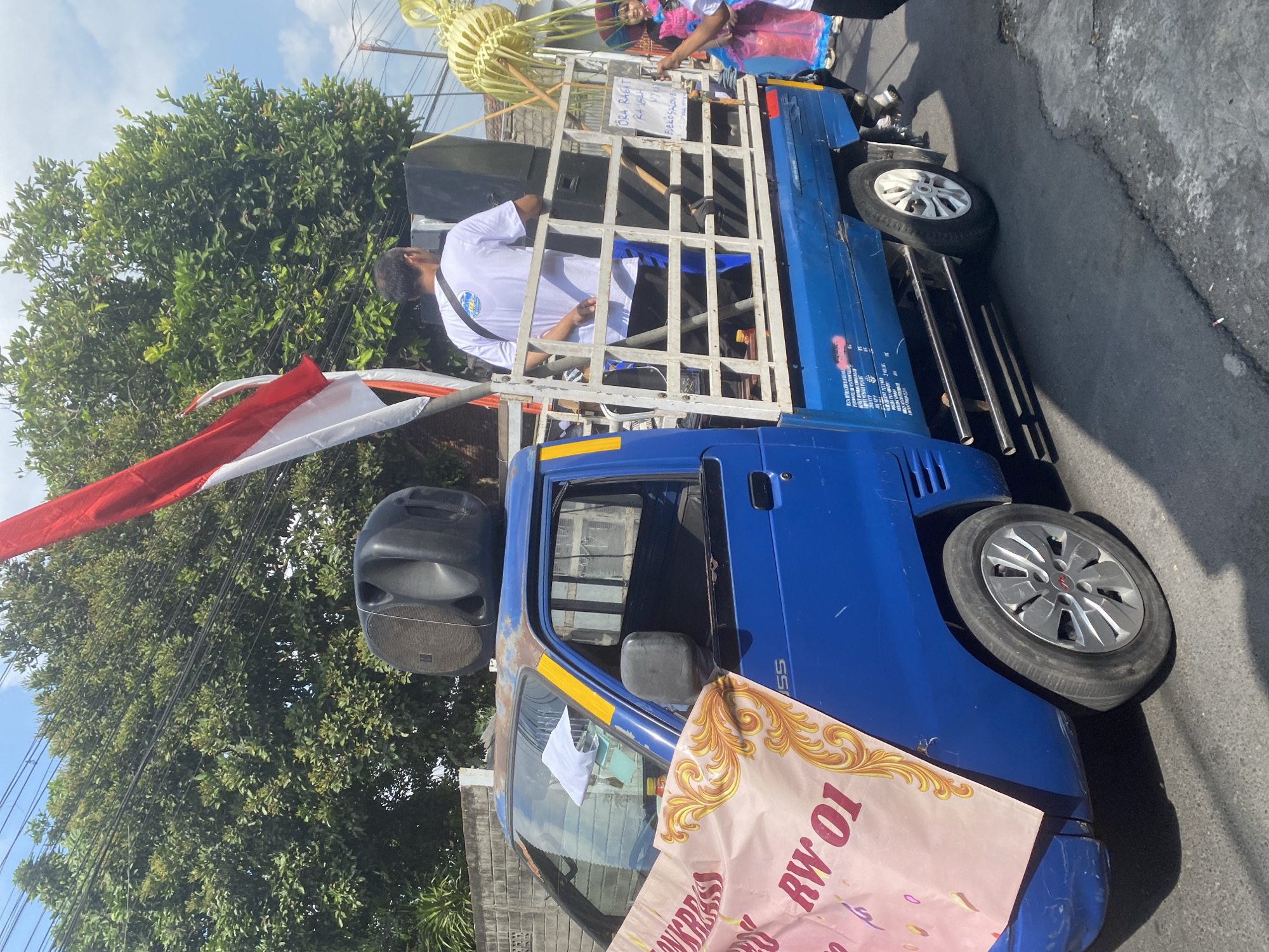 The image depicts a blue pickup truck with a white flatbed being used for a parade or procession in Kediri City, Indonesia. A red and white Indonesian flag is prominently displayed on a pole at the front of the truck bed. Two large black speakers are mounted on the truck bed. A man wearing a white t-shirt with a blue graphic and a dark strap across his chest stands in the truck bed, facing away from the camera. He appears to be holding a microphone or is about to speak. A large pink banner with ornate gold trim and text "RW 01" is attached to the side of the truck's cab. Another banner with text is visible on the side of the truck bed. In the background, there are trees, overhead wires, and a building with a brick wall. To the right, a person in colorful attire, possibly a costume, is partially visible, holding a yellow, straw-like decoration. The truck is driving on a paved road, and the scene appears to be outdoors on a sunny day.