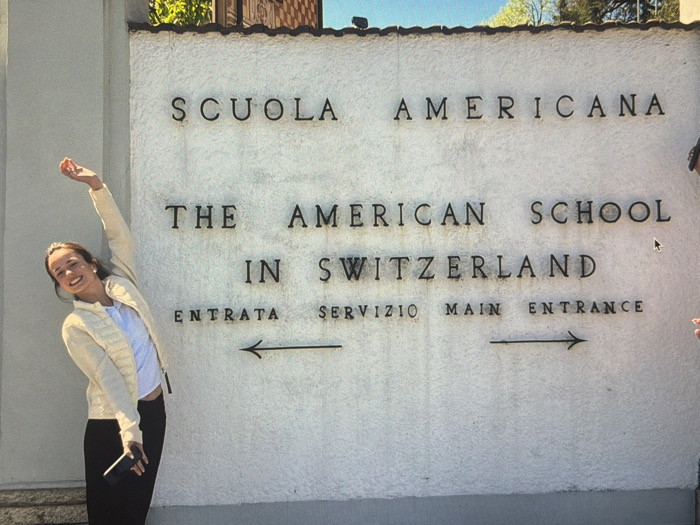 A young woman poses next to a sign that reads "SCUOLA AMERICANA," "THE AMERICAN SCHOOL IN SWITZERLAND," and "ENTRATA SERVIZIO MAIN ENTRANCE," with arrows pointing left and right. She is smiling with her arm raised and holds a phone in her other hand. She is wearing a light yellow jacket, white top, and dark pants. The sign is mounted on a textured white wall.