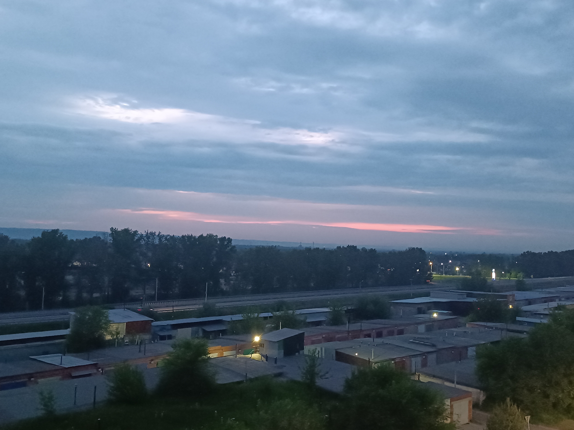 The image shows a twilight scene over a collection of low-rise buildings, likely garages or storage units, in the city of Sosnovoborsk, Russia. The sky is filled with scattered clouds, with a soft pinkish hue near the horizon indicating the fading light of sunset or the approaching dawn. A dense line of trees forms a dark backdrop behind the buildings, with distant lights suggesting further urban development or infrastructure. The foreground consists of more low-lying structures and green foliage, possibly trees or bushes. The overall impression is one of a quiet, possibly industrial or residential outskirts area, captured during a transition period of the day. There are no visible people in the frame.