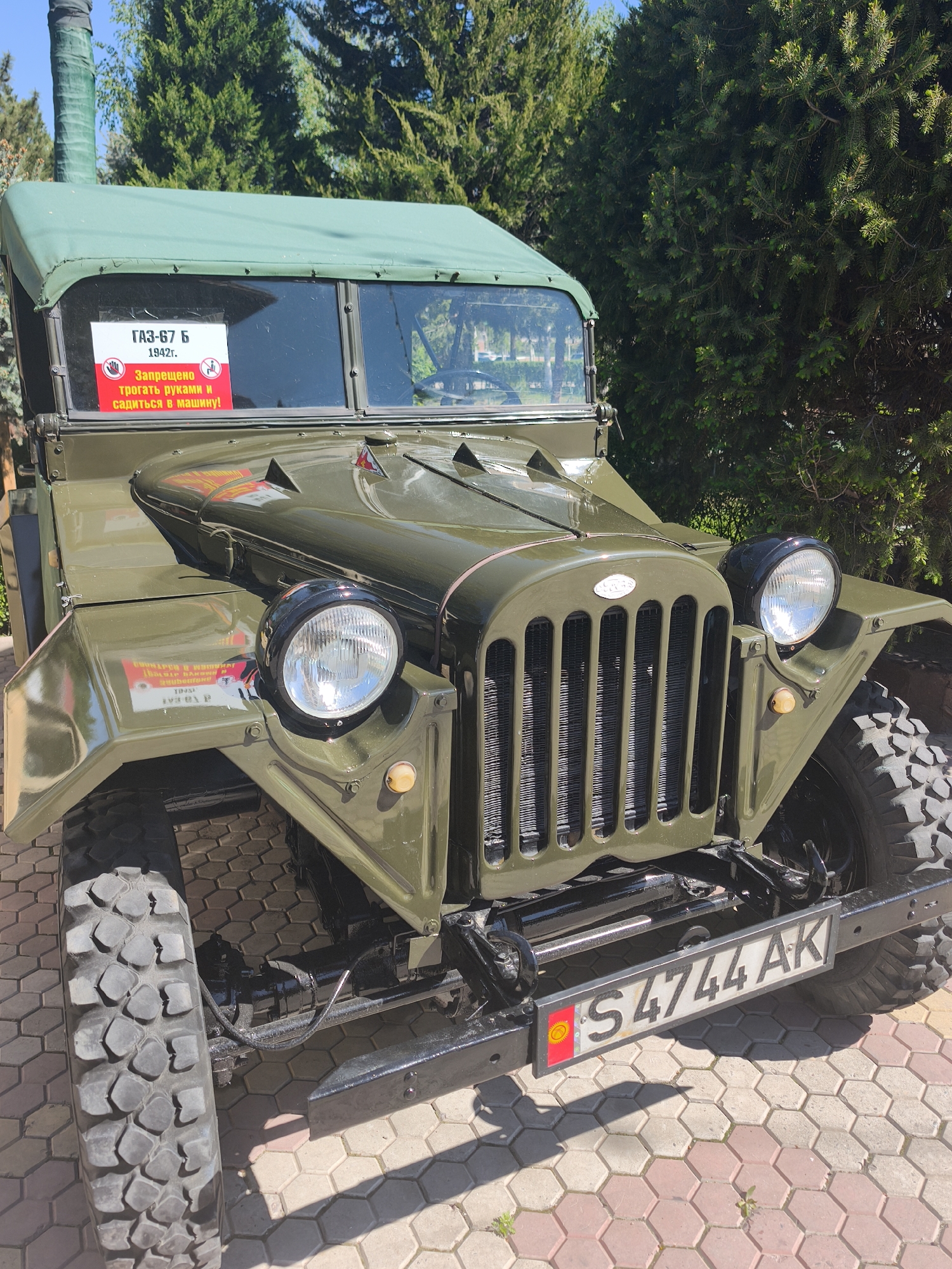 The image captures a vintage military vehicle, specifically a GAZ-67 B from 1942, displayed outdoors. The vehicle is painted in olive green and features distinctive design elements such as large, rounded headlights, a vertical bar grille, and rugged tires. A sign in Russian is affixed to the windshield, prohibiting touching or entering the vehicle. The license plate, marked "S4744 AK," indicates registration in Kyrgyzstan. The setting appears to be Kenbulun, Kyrgyzstan, based on the license plate, with trees providing a backdrop. The vehicle is parked on a paved surface, suggesting a display area or museum.