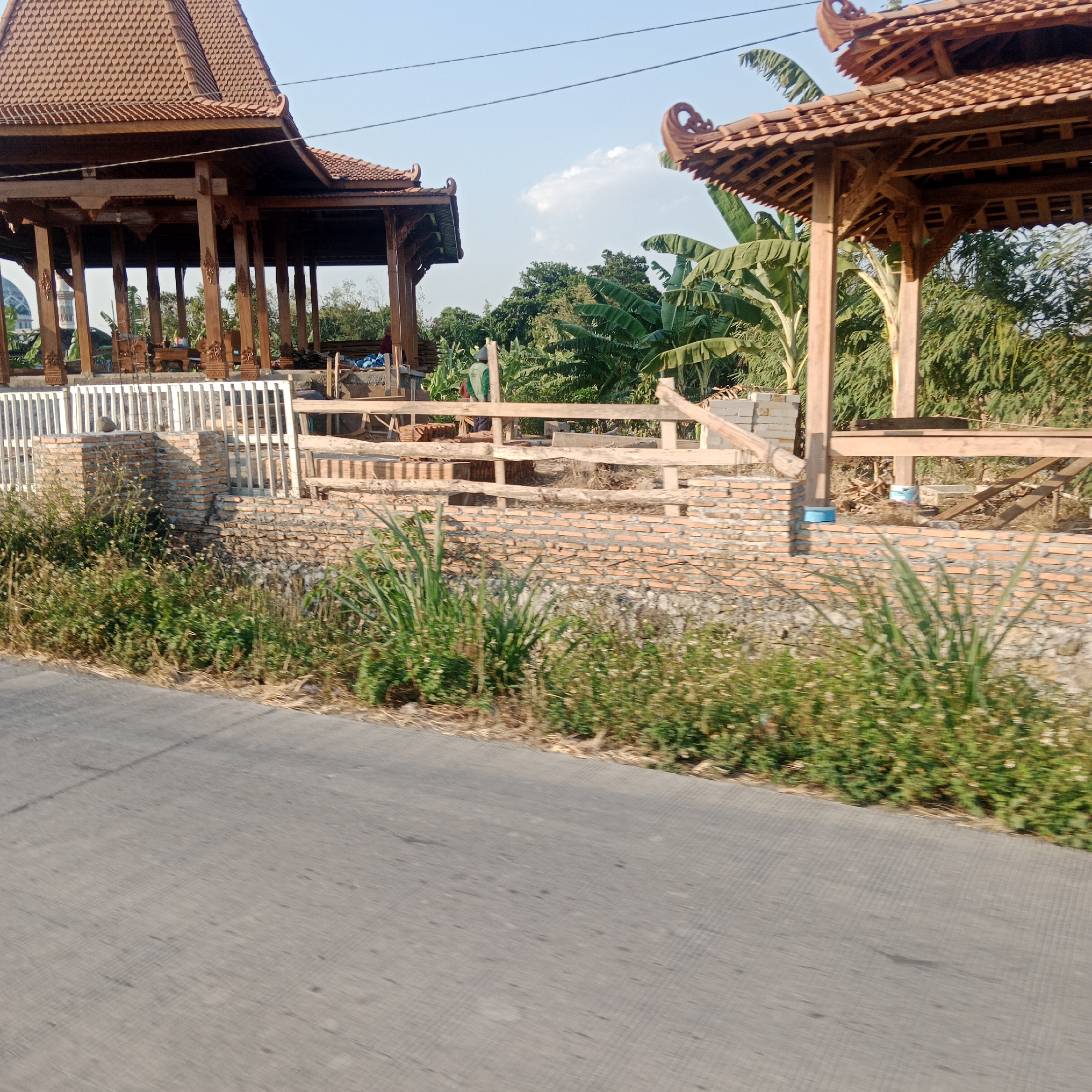 The image depicts an outdoor scene in Jatipecaron, Indonesia, featuring traditional wooden gazebos under construction. The gazebos have ornate, tiled roofs and are supported by carved wooden pillars. In the foreground, a brick wall is visible, partially covered by lush green vegetation and wildflowers. To the left, a white picket fence lines a raised area. The background shows dense greenery, including banana trees and other foliage, under a clear blue sky. A paved road runs along the bottom of the image, suggesting a roadside view. The construction appears to be in its early stages, with exposed wooden beams and building materials present. A person in a green shirt and a hat is visible near the construction area, possibly overseeing the work.