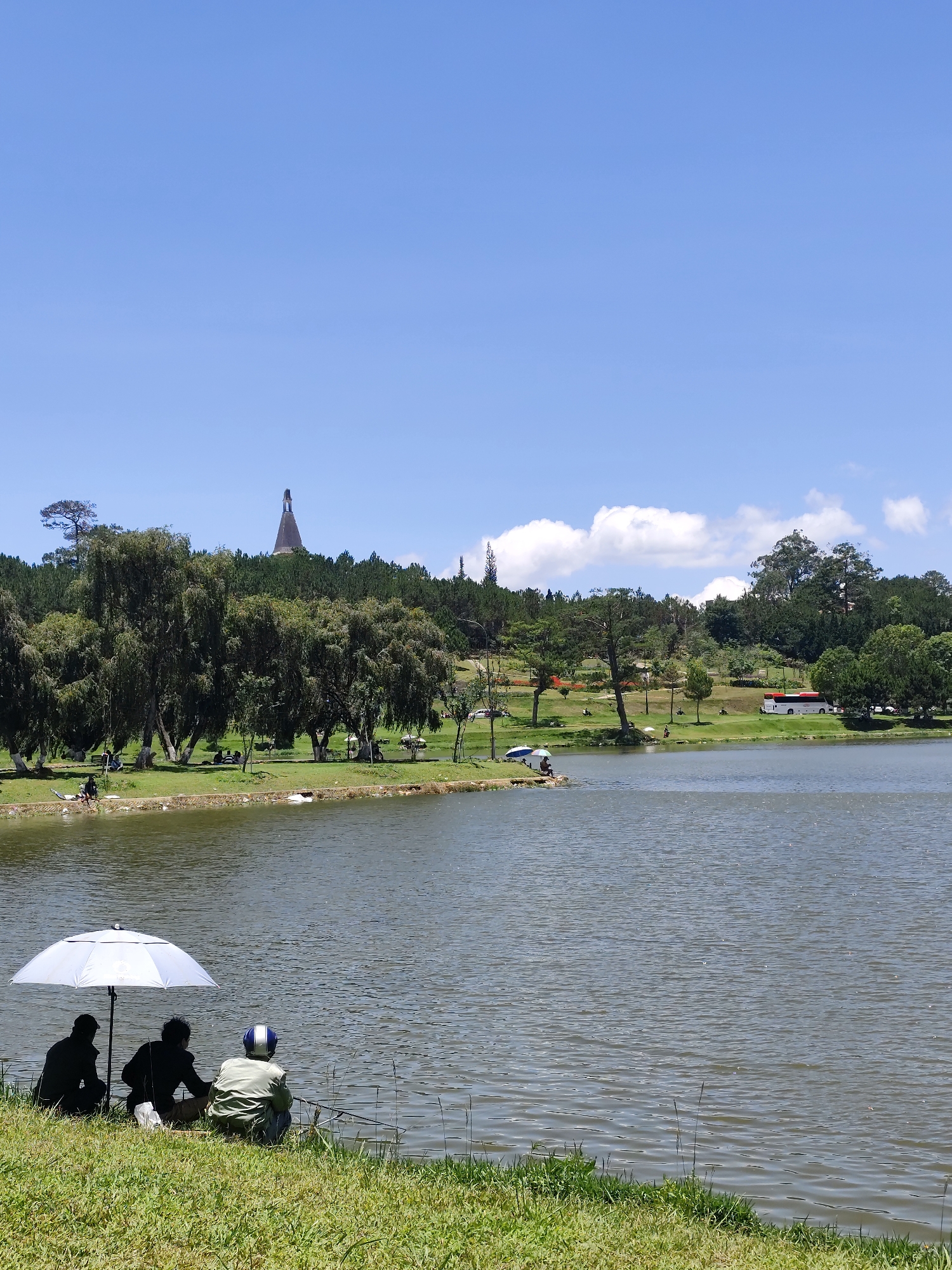 The image shows a lake in Da Lat, Vietnam, likely Xuan Huong Lake, on a sunny day. The sky is mostly clear blue. In the foreground, three people are sitting on the grassy bank of the lake, fishing. A white umbrella shades them. One person wears a motorcycle helmet. In the background, the opposite shore is visible with trees, a grassy park area with a few scattered people, and a tall, conically-shaped structure on a hill. A single low cloud is present.