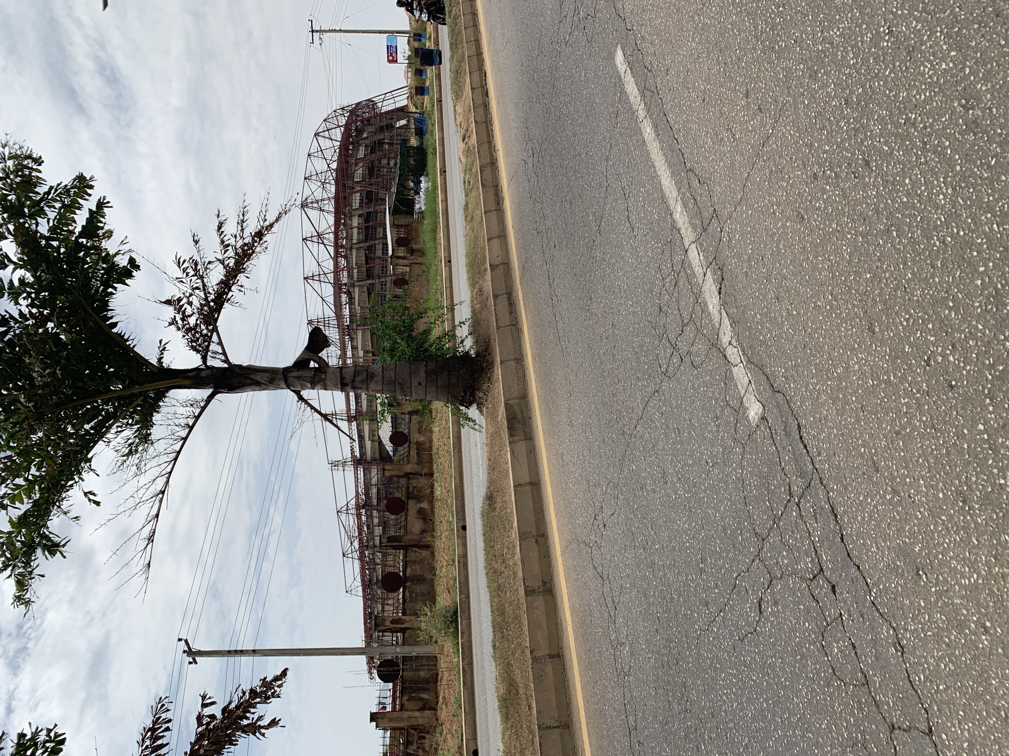 The image depicts a section of roadway in close proximity to the New Stadium in Nigeria. The road is asphalt, with a painted white line marking the lane. Cracks are visible in the pavement.
Adjacent to the road is a partially constructed building featuring scaffolding and structural supports. A tree is present, partially obscuring the view of the building. Utility poles with power lines are also visible. The sky is overcast with cloudy conditions.